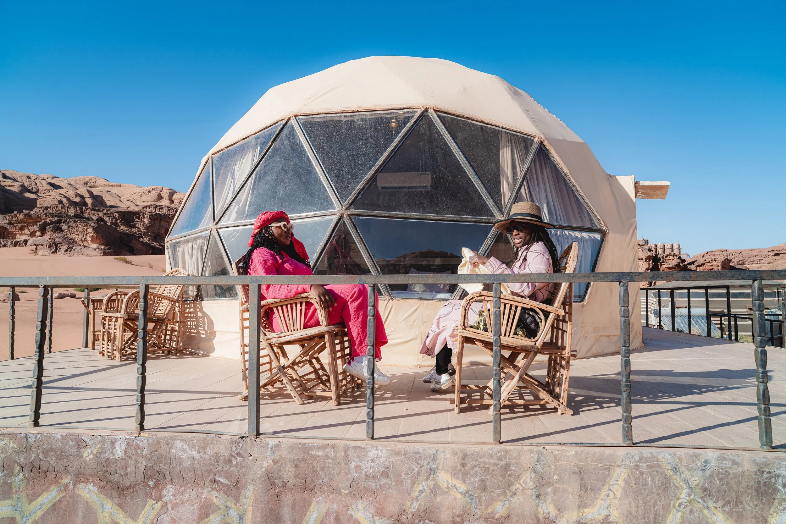 Two women sitting on wooden chairs outside a geodesic dome in a desert landscape