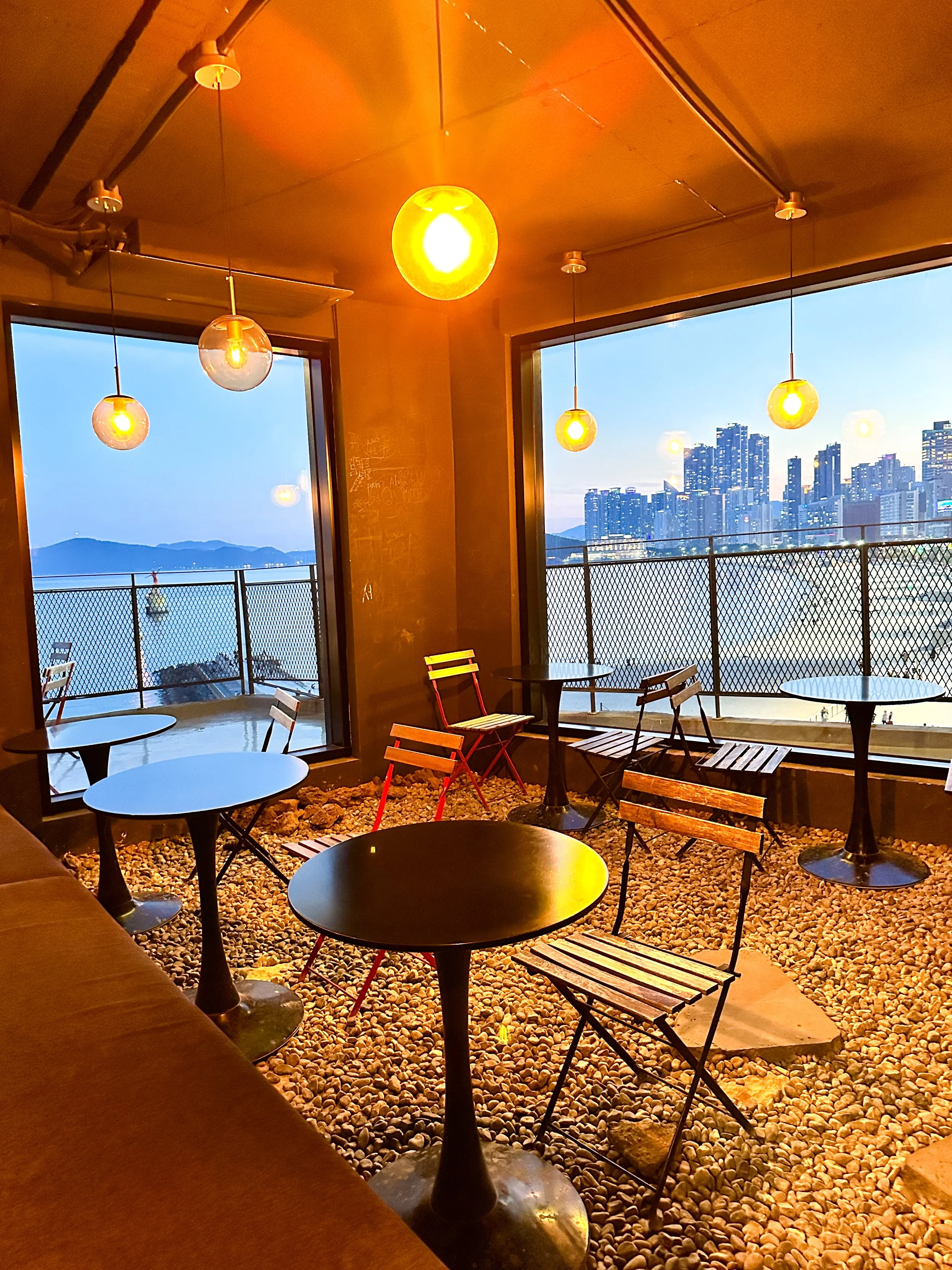 Indoor seating area with small round tables and wooden chairs with a rocky floor, illuminated by warm hanging lights. Large windows offer a view of the city skyline and waterfront at dusk.
