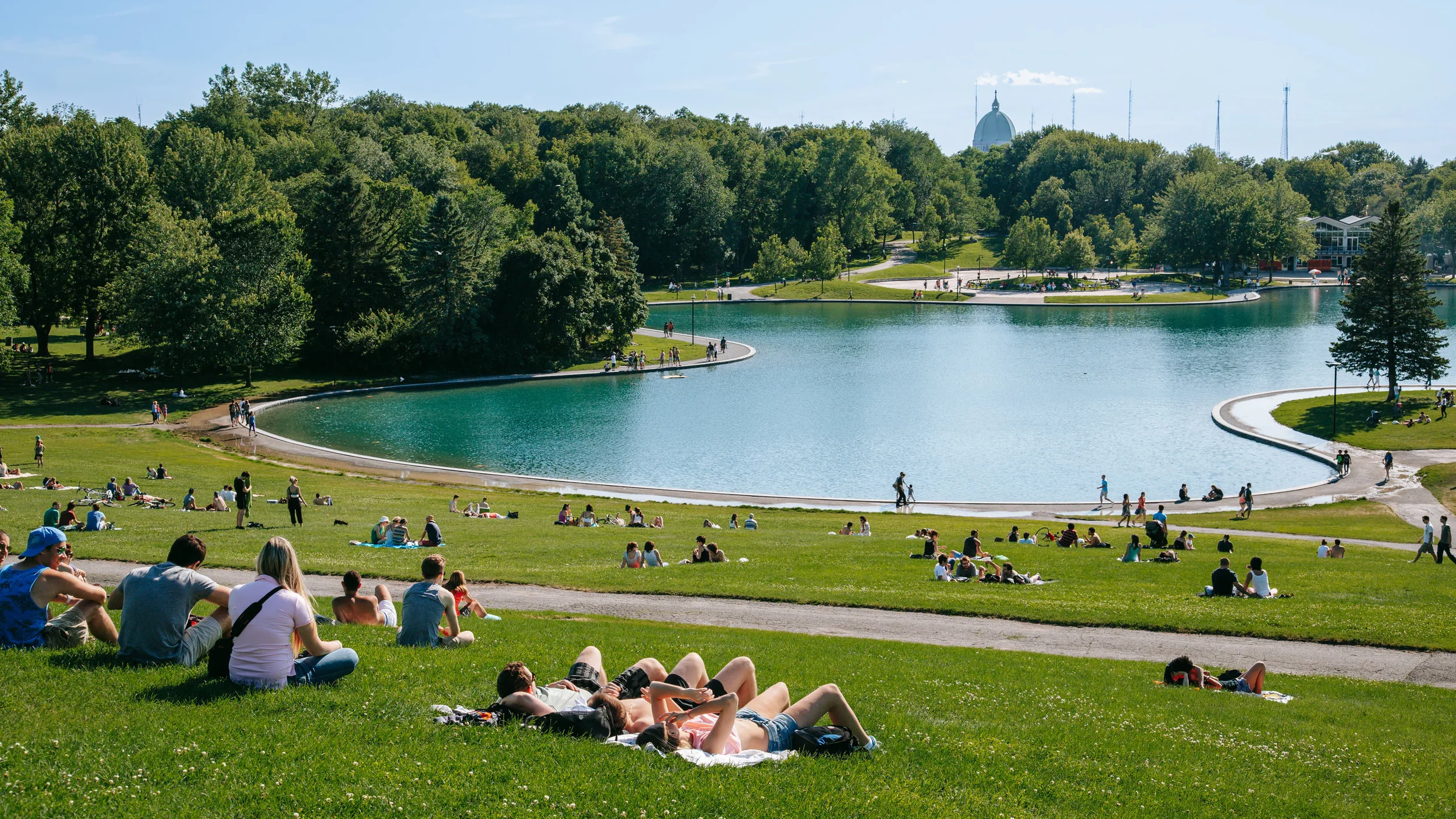 People relaxing on grass near a lake in a park with trees and a city skyline in the background.