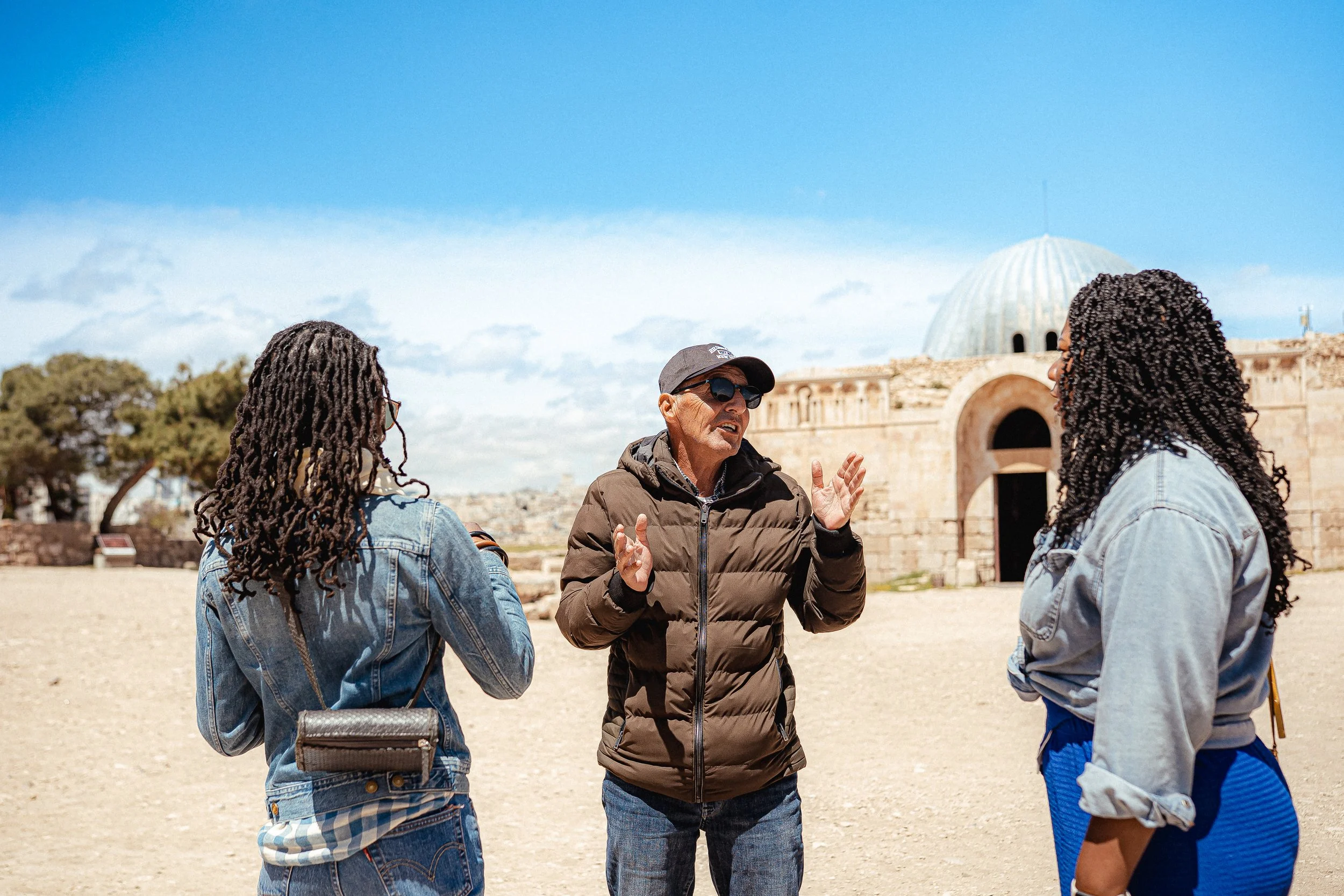 Tour guide explaining to two women at historical site with ancient ruins and a dome in the background.