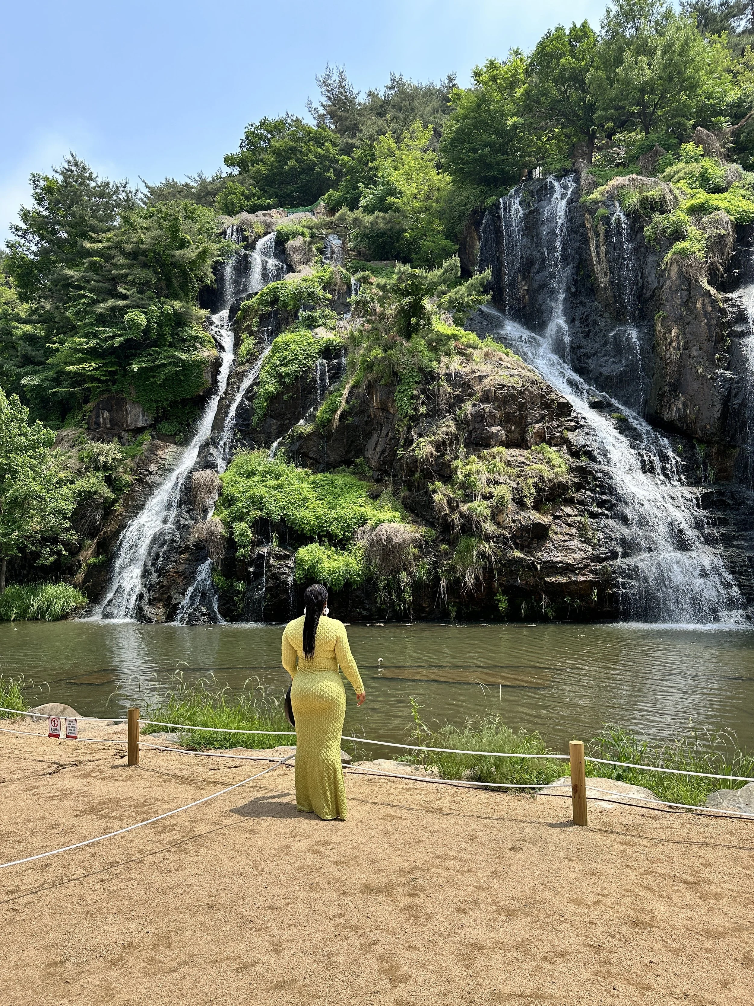 A woman in a yellow dress standing by a pond and observing multiple waterfalls cascading down a rocky hillside surrounded by lush green trees.