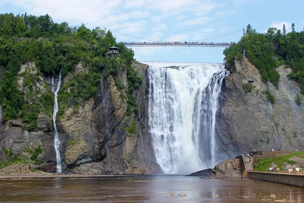 Niagara Falls with a bridge overhead, surrounded by lush green trees, and a viewing area with visitors.