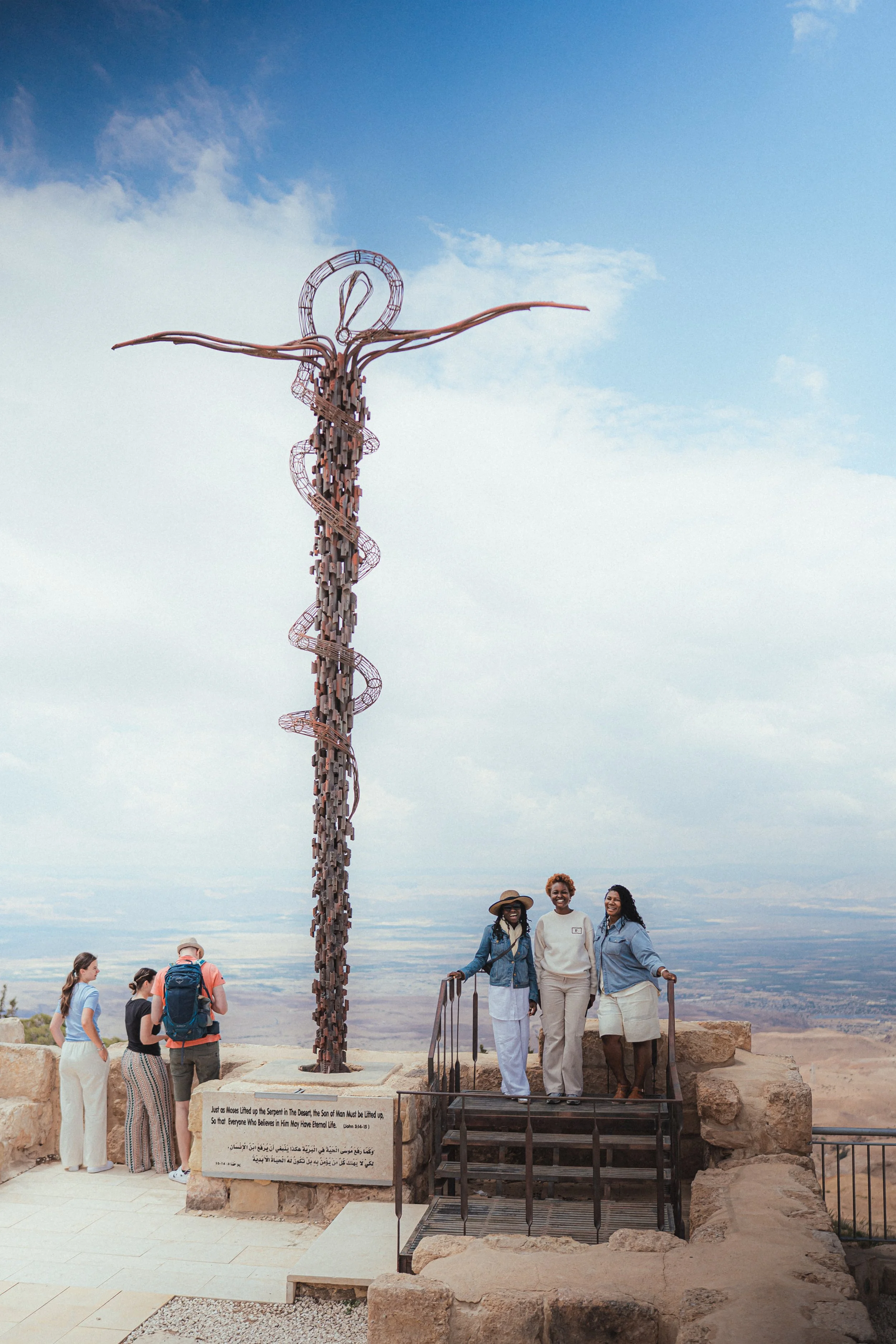 People taking photos beside a tall metal sculpture of a serpent winding around a staff, set on a raised platform with a scenic desert landscape in the background.