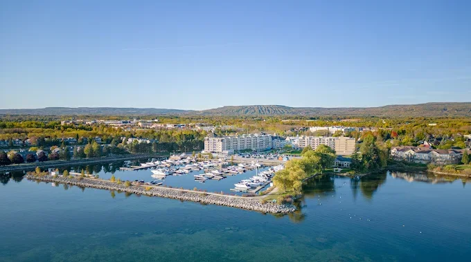 Aerial view of a marina with boats, surrounded by water and buildings, with a suburban area and hills in the background under a clear blue sky.