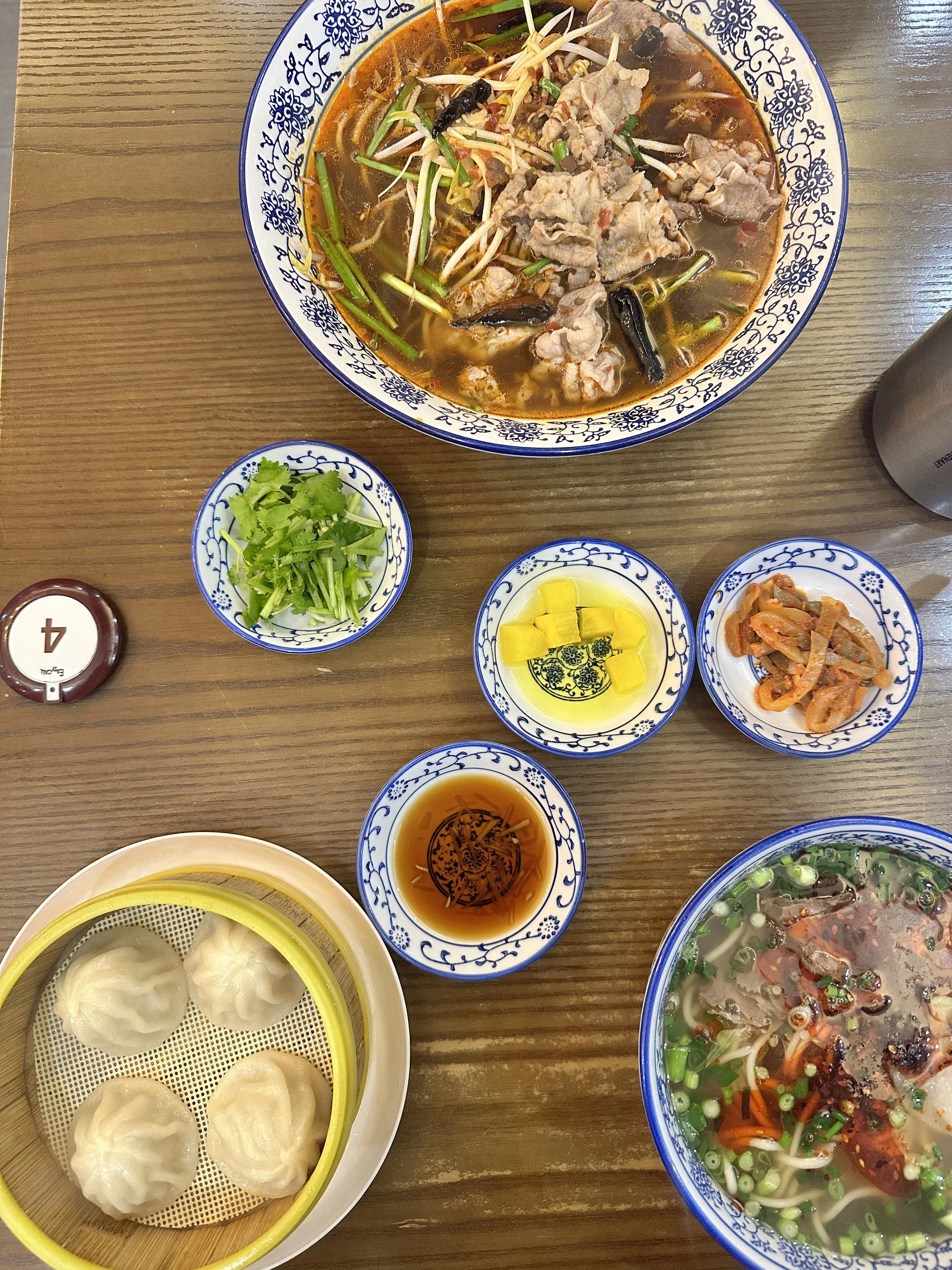 A table with Asian dishes including a bowl of spicy soup with meat and vegetables, a bowl of clear soup with sliced meat and green onions, a small basket of steamed dumplings, and small bowls of pickled vegetables, greens, and dipping sauces.