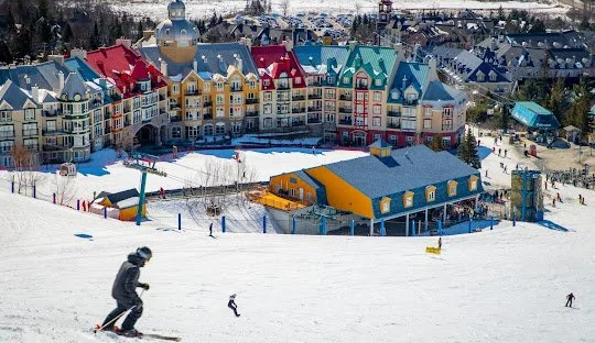 Skiers on snowy slope in front of colorful resort buildings with snow-covered rooftops.