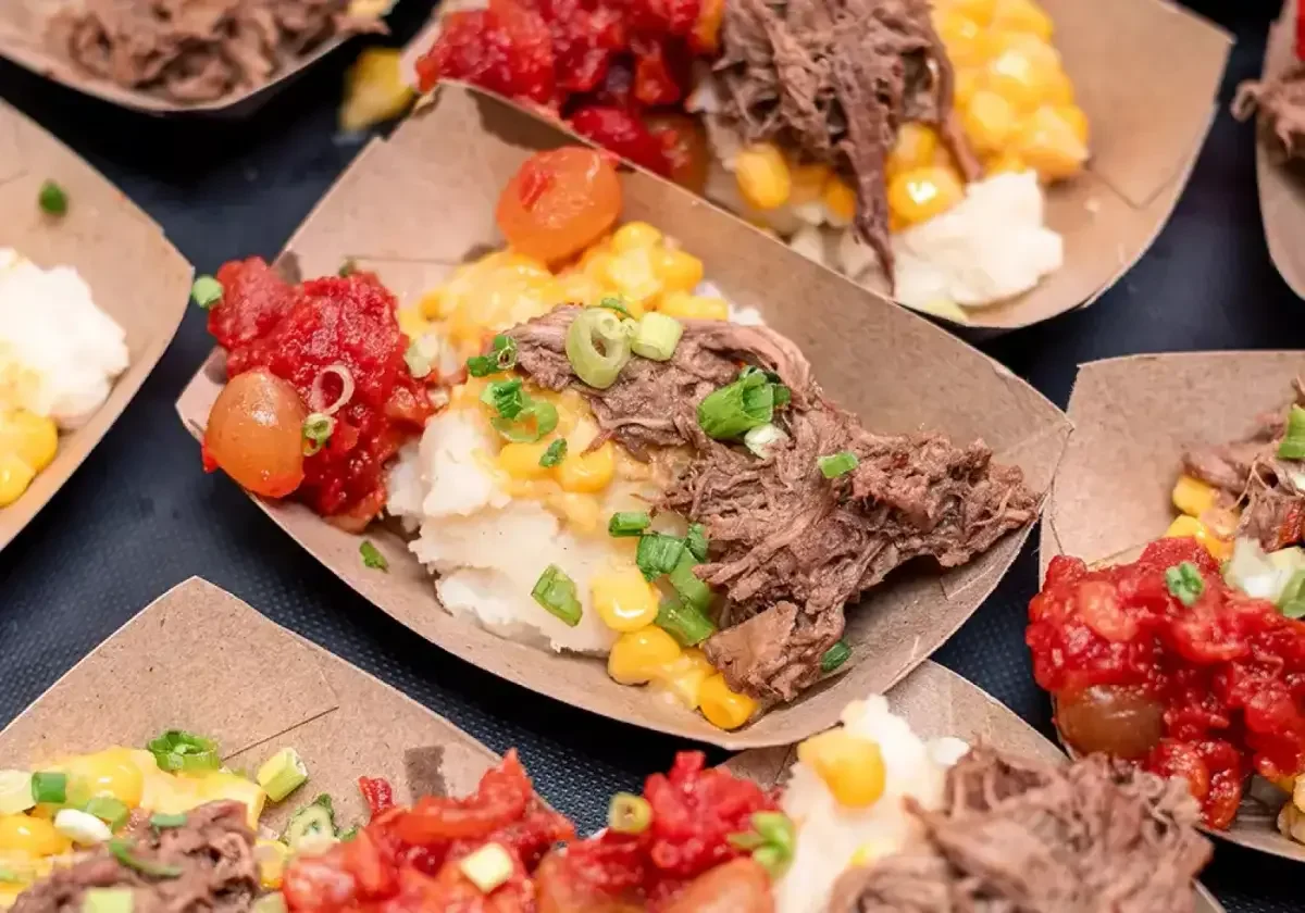Several taco servings with shredded beef, corn, tomato salsa, chopped green onions, and sour cream in small paper trays.