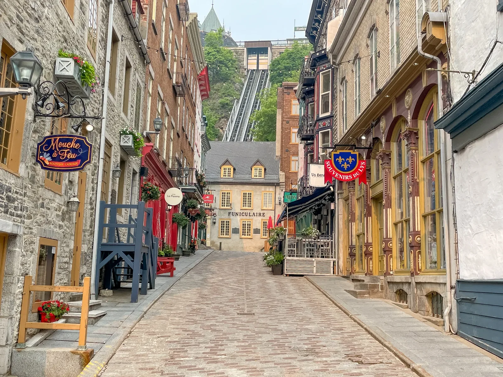 A narrow cobblestone street in a European-style neighborhood with colorful buildings, flower boxes, hanging signs, outdoor seating, and a funicular railway in the background on a hill.
