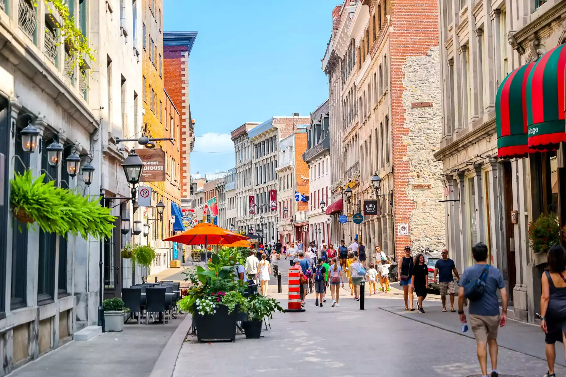 A lively city street with colorful buildings, outdoor cafes with umbrellas, and pedestrians walking on a sunny day.