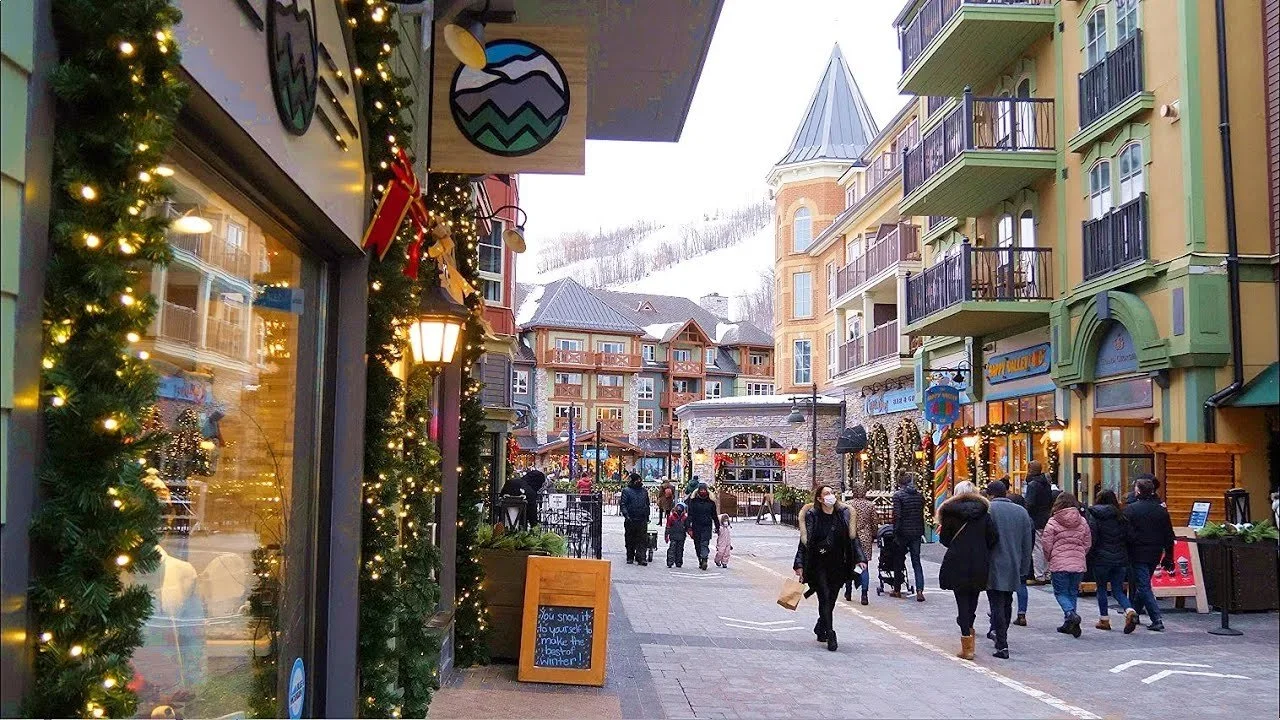 An outdoor shopping area decorated with Christmas lights and garlands. People are walking along the street, some with shopping bags. The buildings have a European architectural style with balconies and colorful facades. Snow is visible on the rooftop