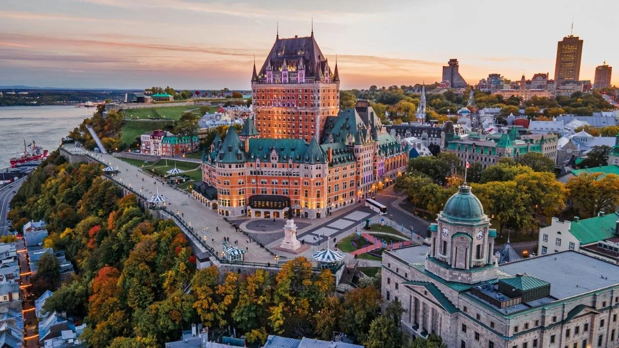 Aerial view of the Château Frontenac hotel in Quebec City during sunset, with colorful autumn trees and the Saint Lawrence River in the background.