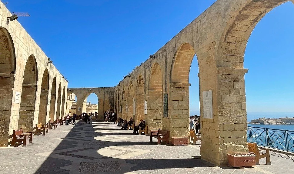 Stone archways along a promenade with benches and people walking, overlooking the sea on a sunny day.