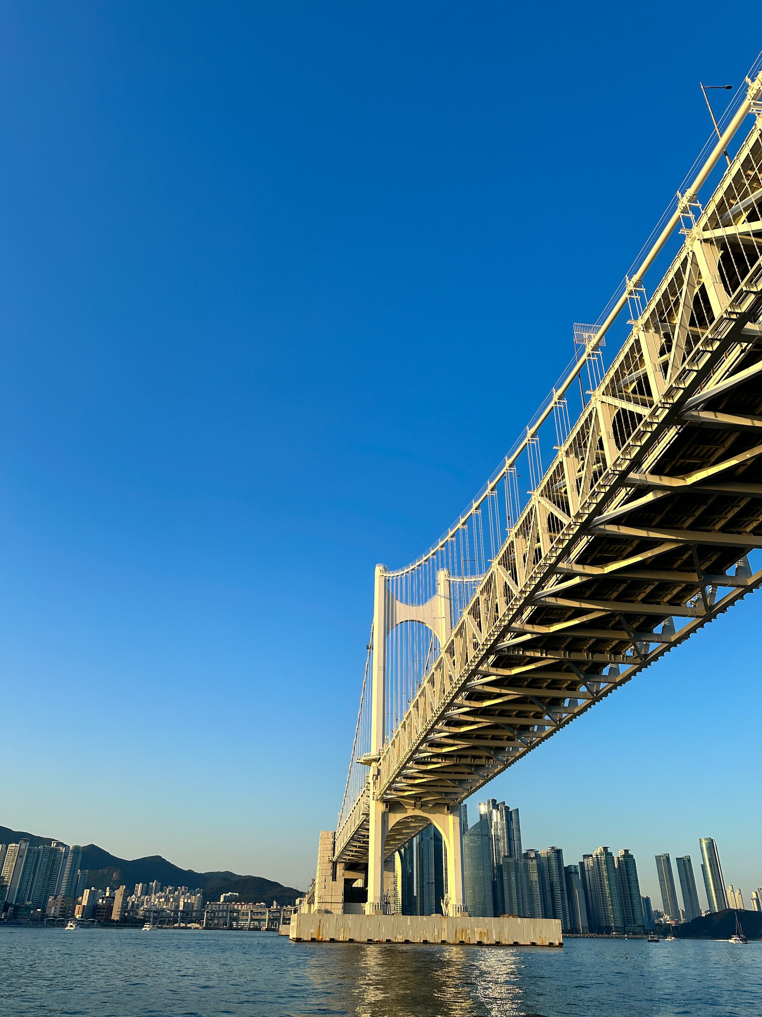 Photo of the Akashi-Kaikyo Bridge in Japan, showing the suspension bridge extending over the water with city buildings and mountains in the background.