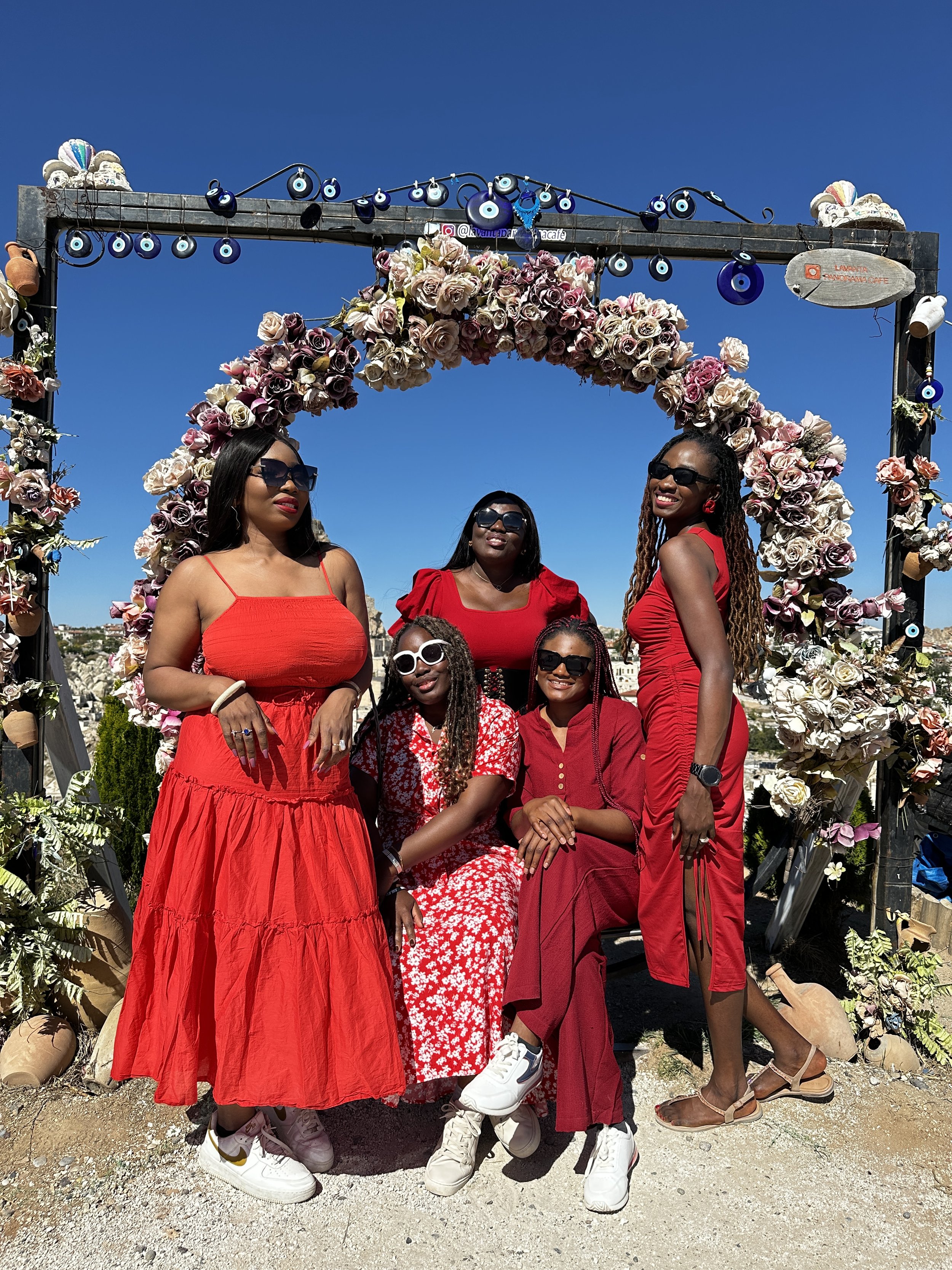 Five women wearing red dresses posing together under a floral arch at what appears to be an outdoor event on a sunny day.
