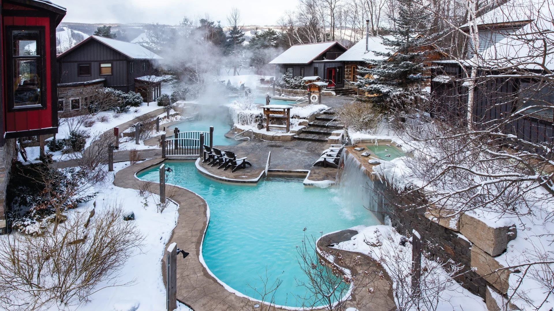 A snowy outdoor hot spring resort with multiple pools, steam rising from the water, surrounded by trees and wooden cabins.