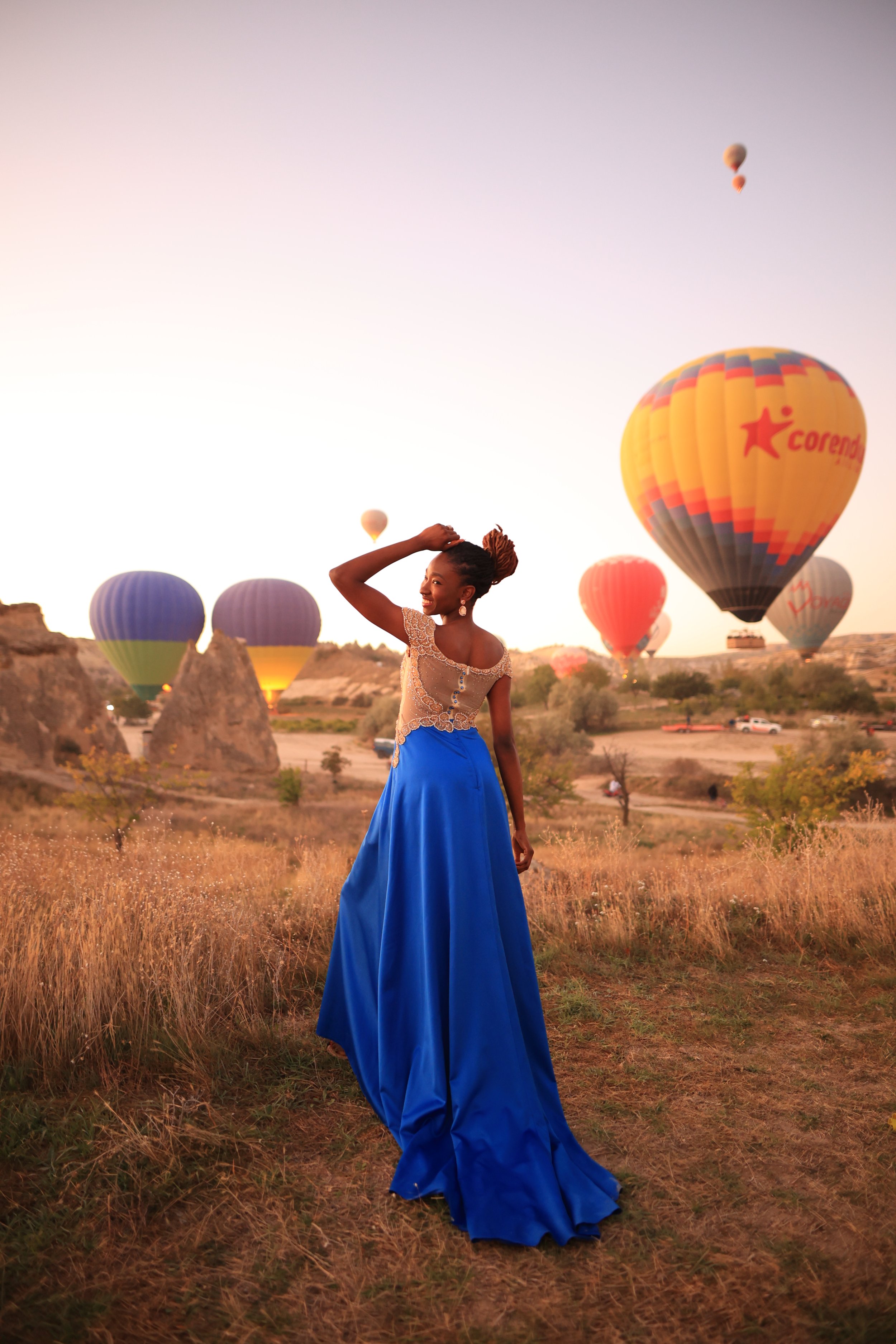 A woman standing in a field at sunset with hot air balloons in the background.