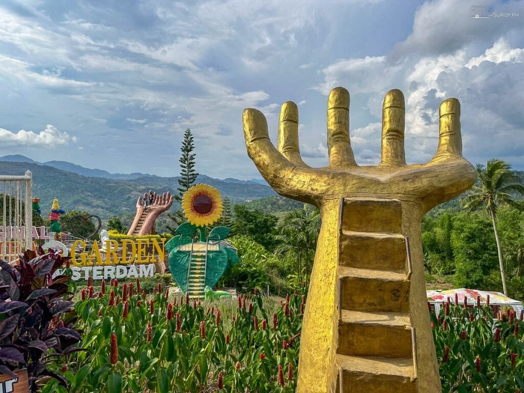 A large golden sculpture of a hand with an open palm, with stairs leading up to the palm, surrounded by lush greenery, colorful flowers, and decorative signs reading 'GARDEN' and 'SVERDAM.' In the background, there's a giant sunflower, a smaller hand sculpture, and a scenic mountain view under a partly cloudy sky.