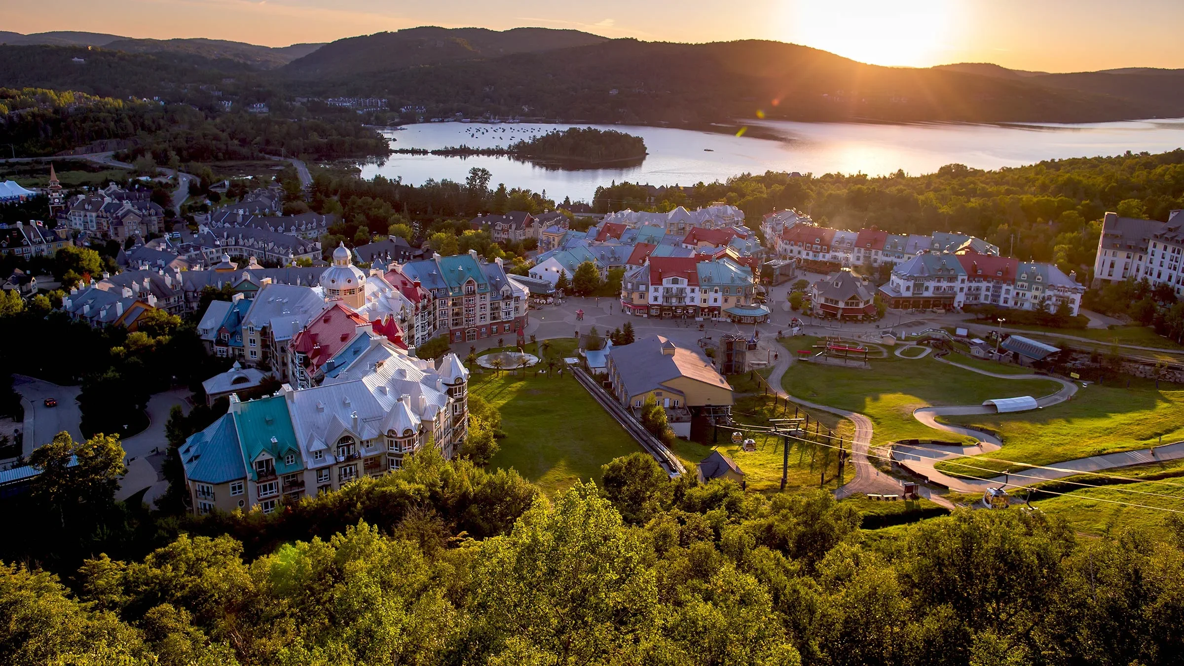 Aerial view of a picturesque town by a lake during sunset, with colorful buildings, green spaces, and surrounding hills.
