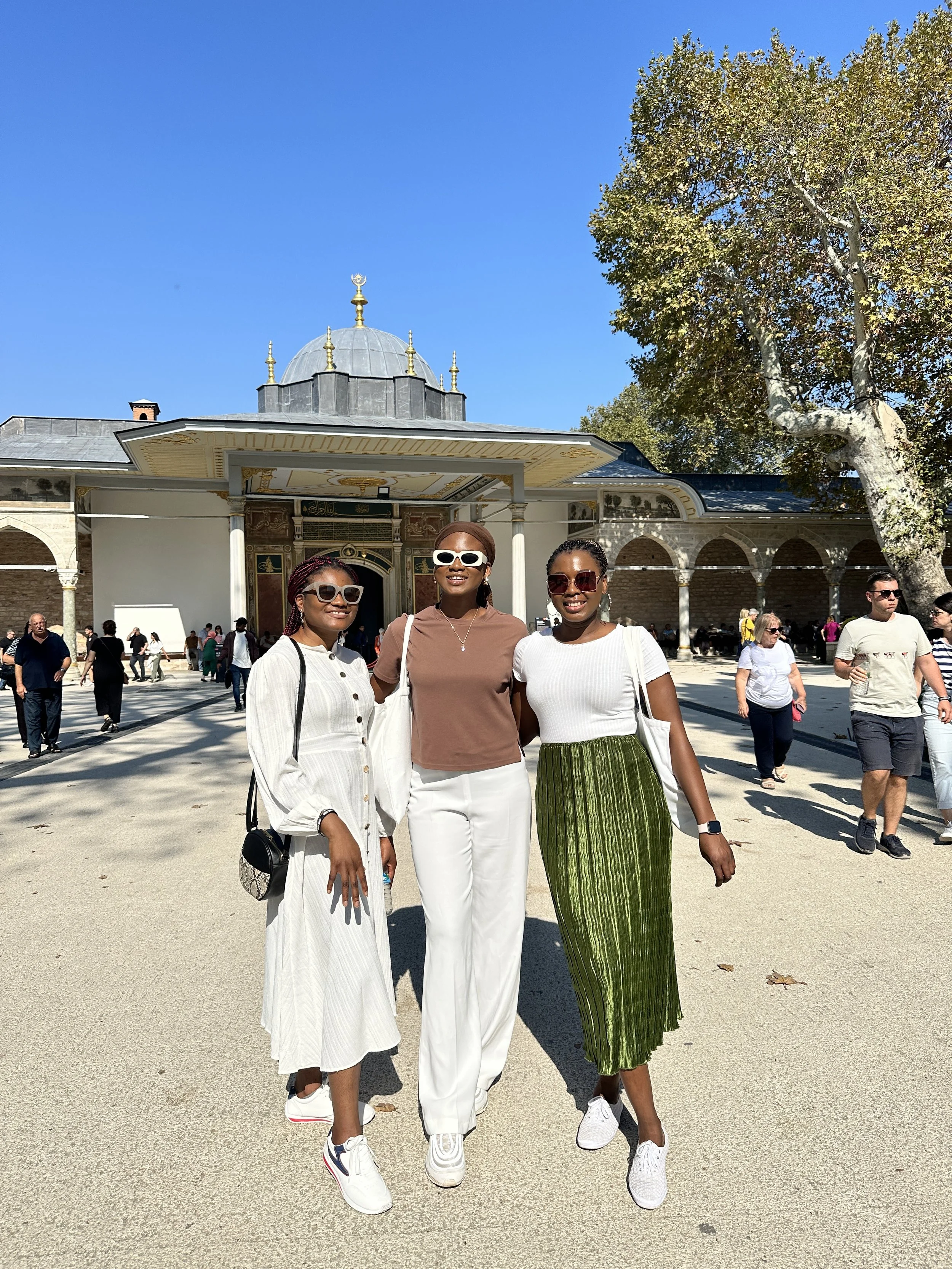 Three women in casual clothing standing together outdoors in front of a historical building with a dome under a clear blue sky.