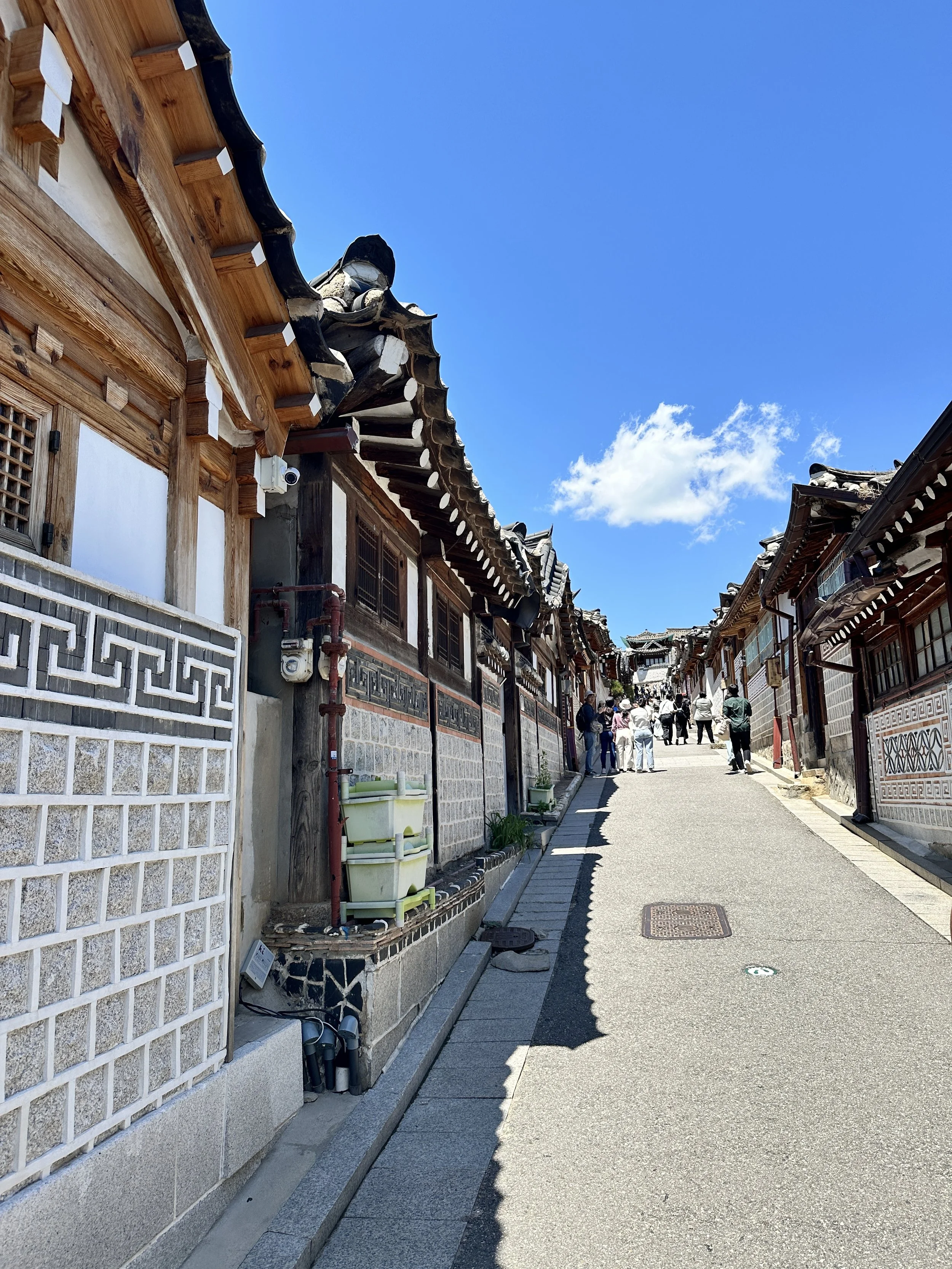 Traditional Korean hanok houses with tiled roofs lining a narrow street on a sunny day with a bright blue sky and a few clouds.