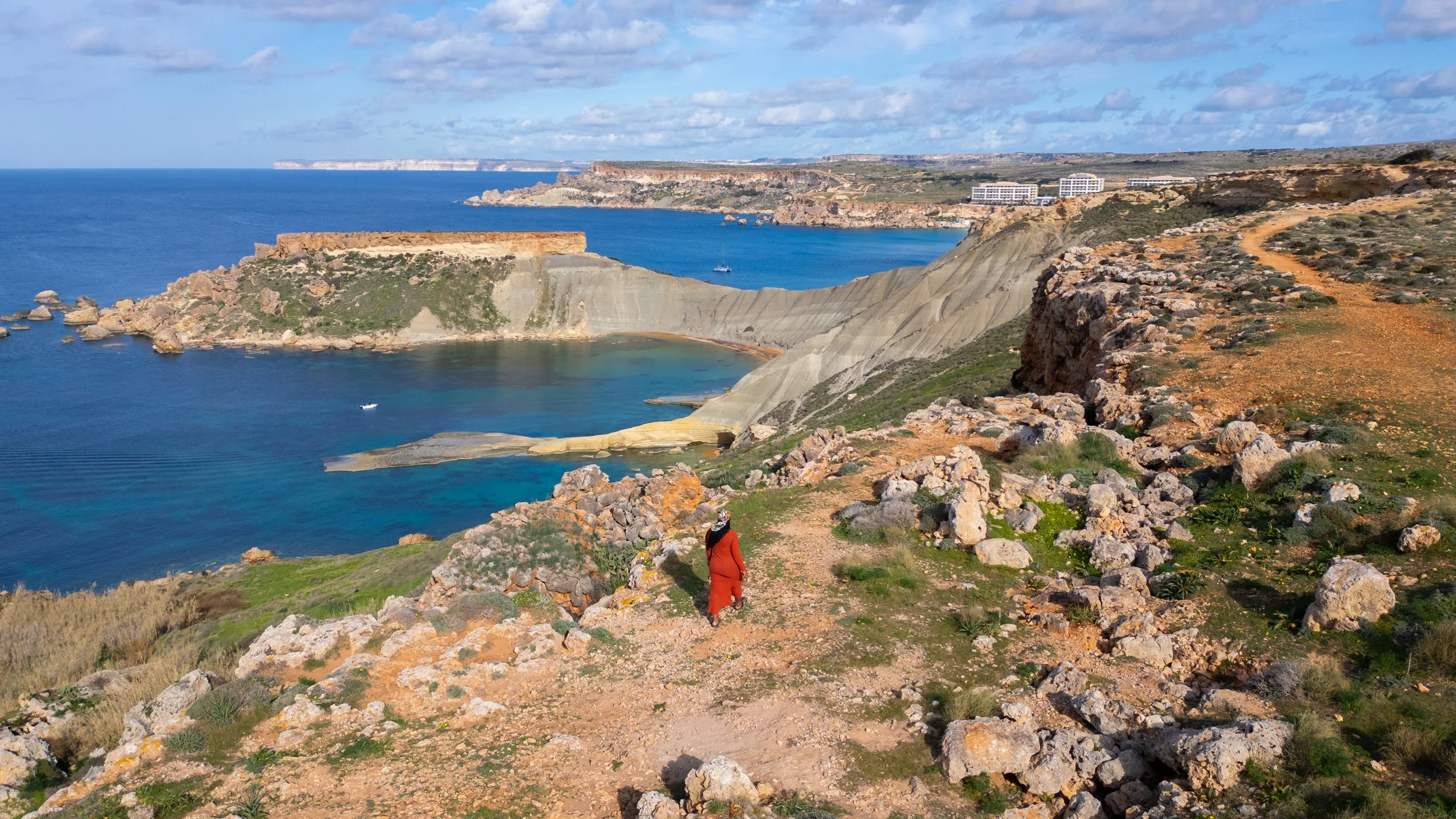 A person walking on a dirt trail along a rocky coastline with cliffs and vivid blue water, overlooking a bay with boats and distant buildings, under a partly cloudy sky.