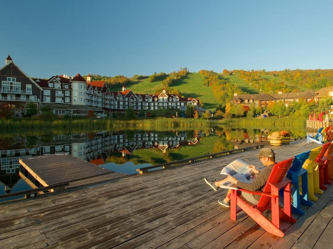 A person sitting on a red chair on a wooden dock by a lake, reading a newspaper, with colorful chairs nearby, overlooking buildings and green hills reflected in the water at sunset.