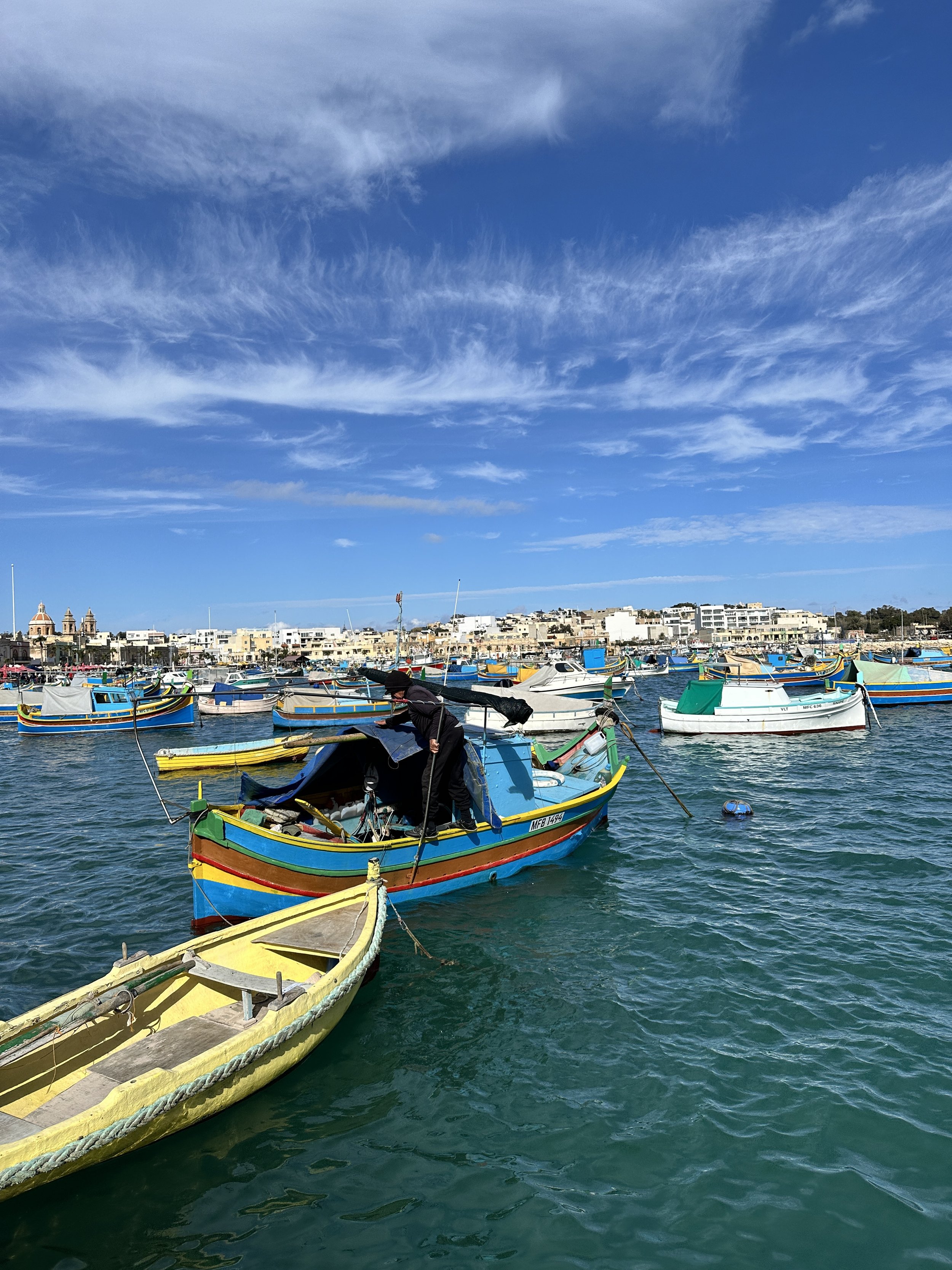 Colorful boats docked in a marina on a clear day with blue sky and wispy clouds, and buildings in the background.