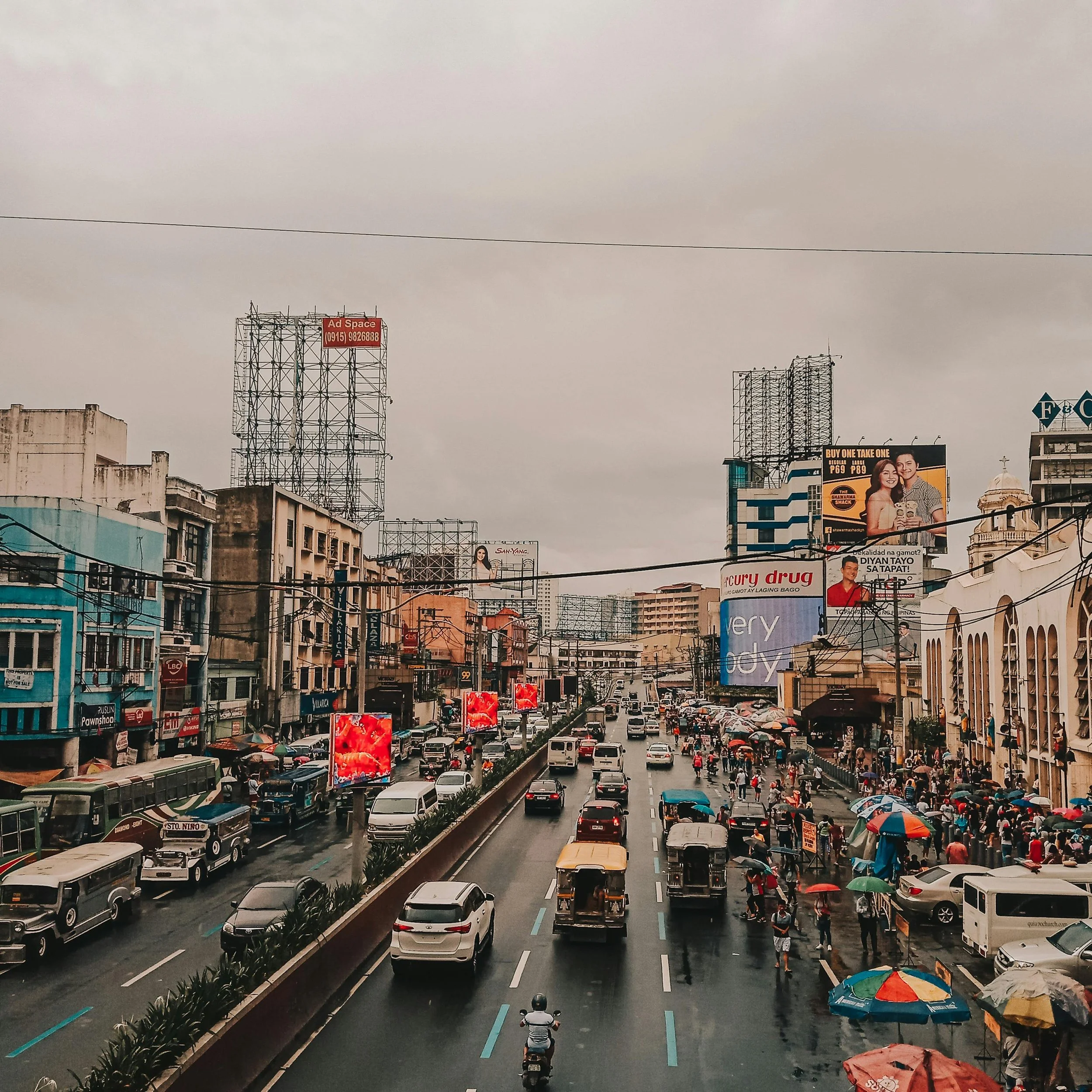 Urban street scene during rainy day with cars, motorcycles, and pedestrians with umbrellas, high-rise buildings, and billboards under cloudy sky.