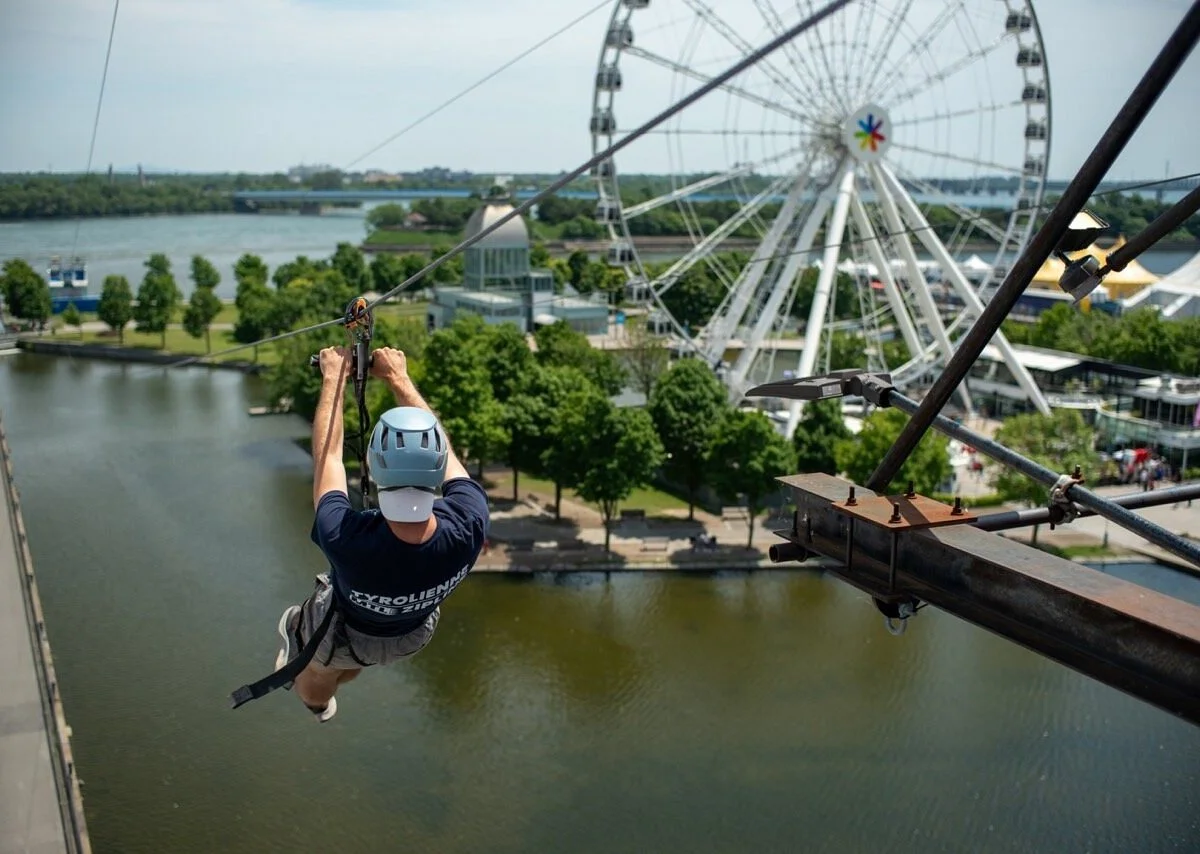 Person wearing a helmet and harness, zip lining over a river at an amusement park with a large Ferris wheel in the background on a partly cloudy day.