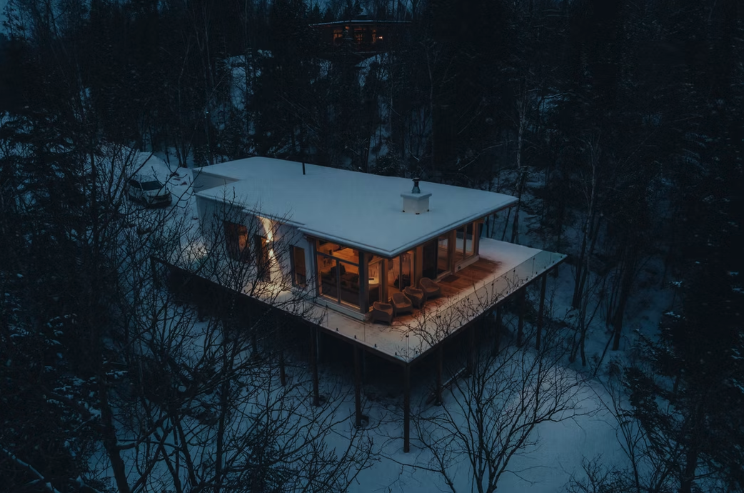 A modern house with a flat roof surrounded by snow, with a large deck and warm interior lights visible through large windows, in a wooded area during winter at dusk.