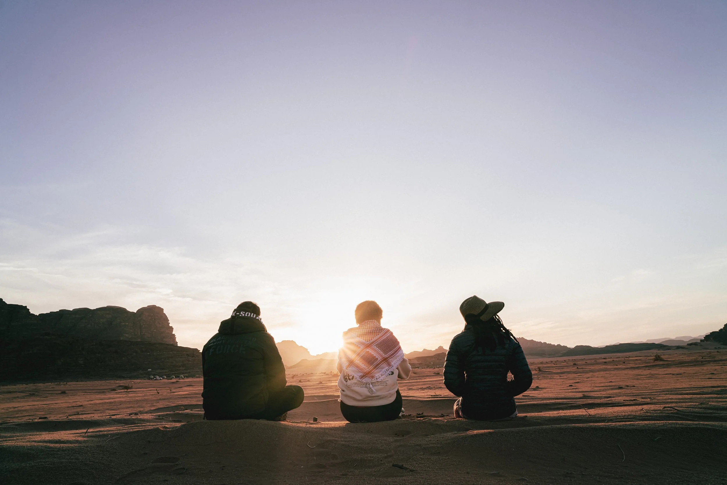 Three people sitting on the desert ground, watching the sunset over mountains.