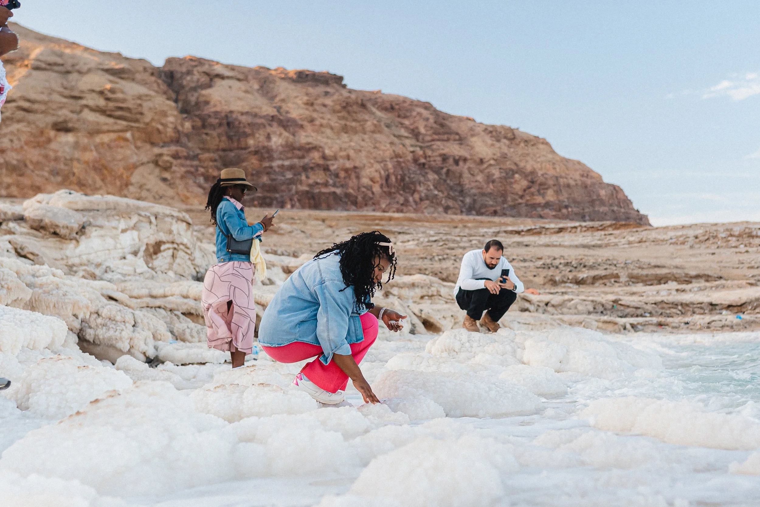 Three people exploring salt formations on a shoreline with mountains in the background, two women and one man, all using their phones.