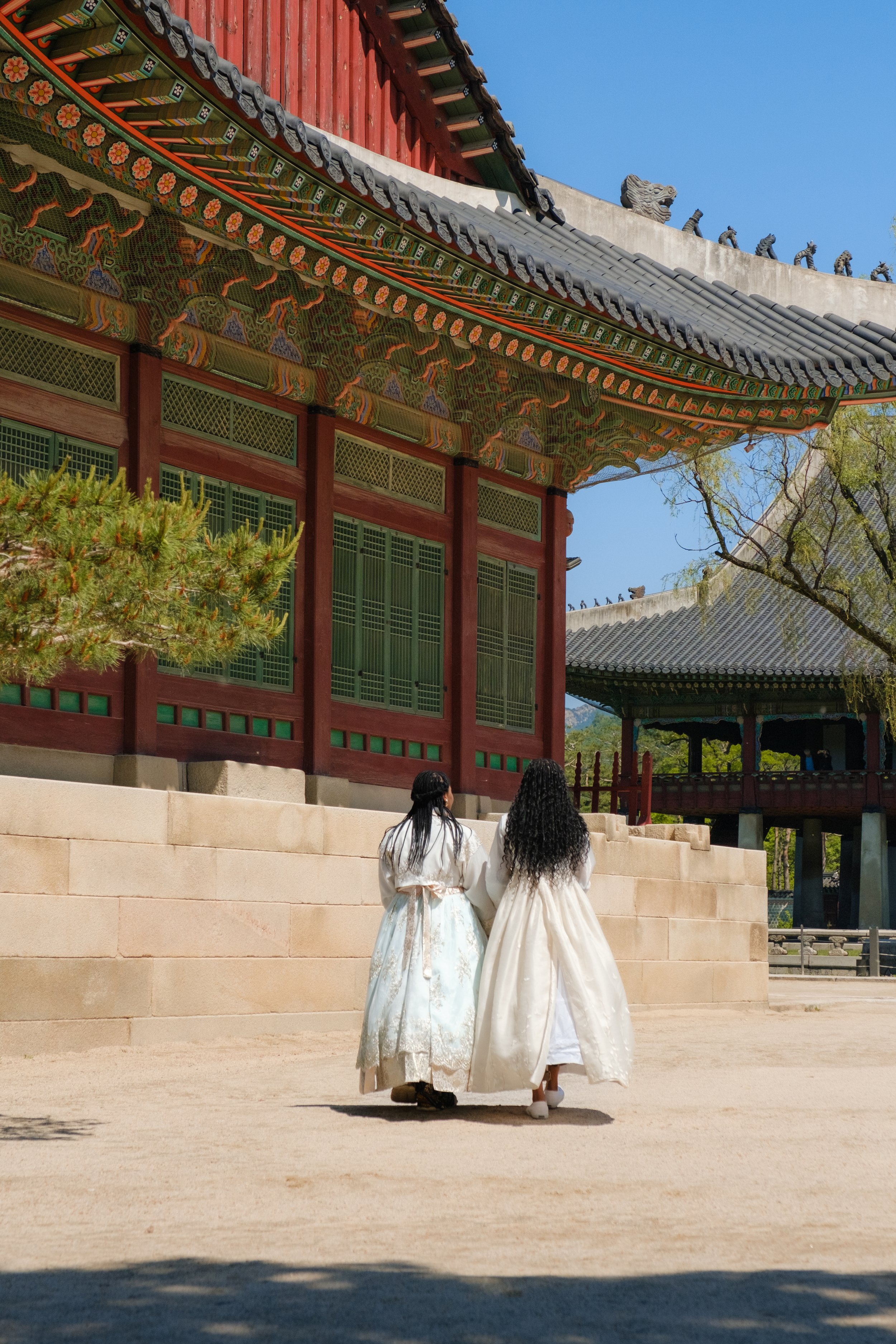 Two women walking towards a traditional Korean temple, wearing historical Hanbok dresses, surrounded by trees and a clear blue sky.
