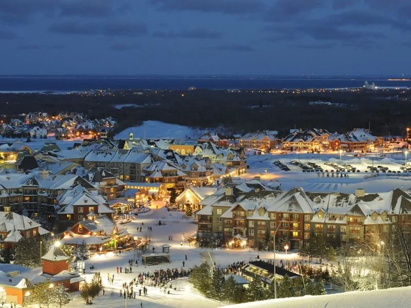 Snow-covered resort town at night with lit buildings, outdoor ice skating rink, and people enjoying winter activities.