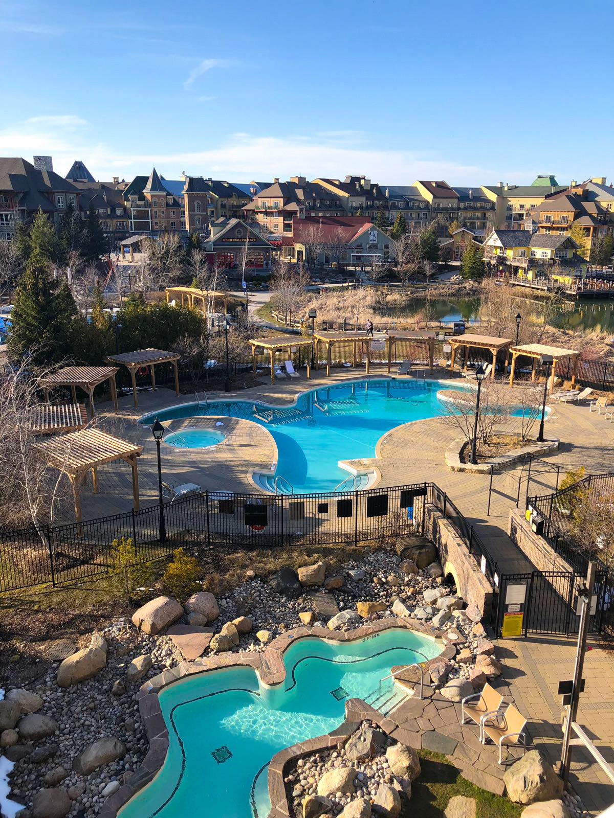Outdoor swimming pools and hot tub area in a residential complex with a pond and buildings in the background.