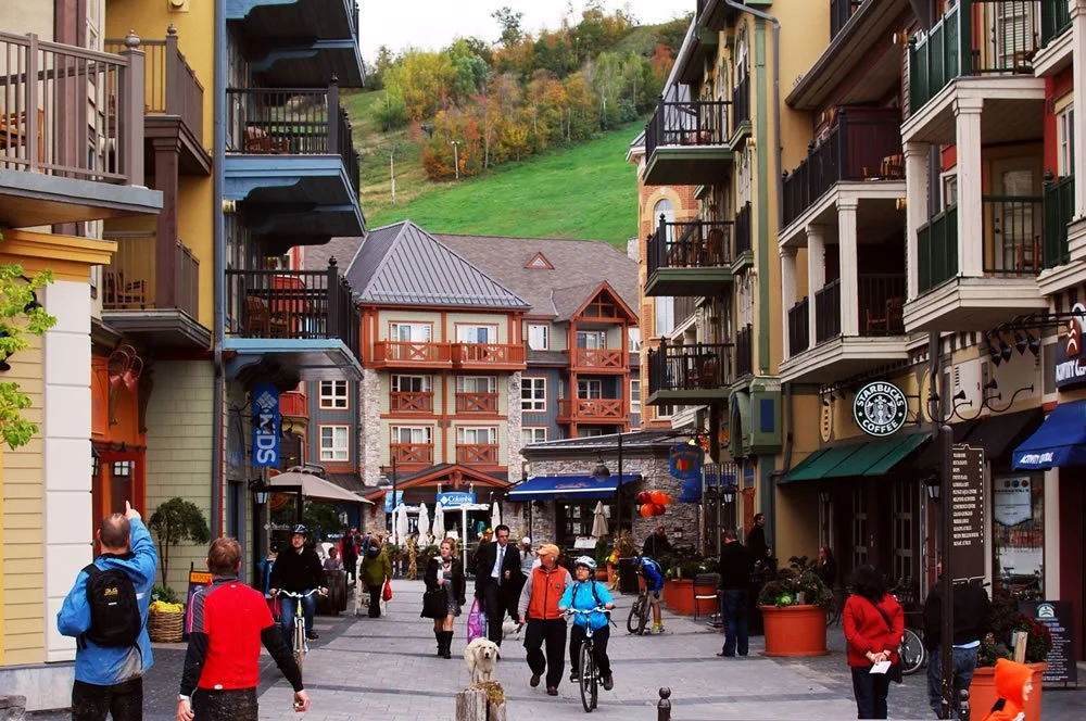 People walking and cycling in a shopping district with colorful buildings and a backdrop of green hills.