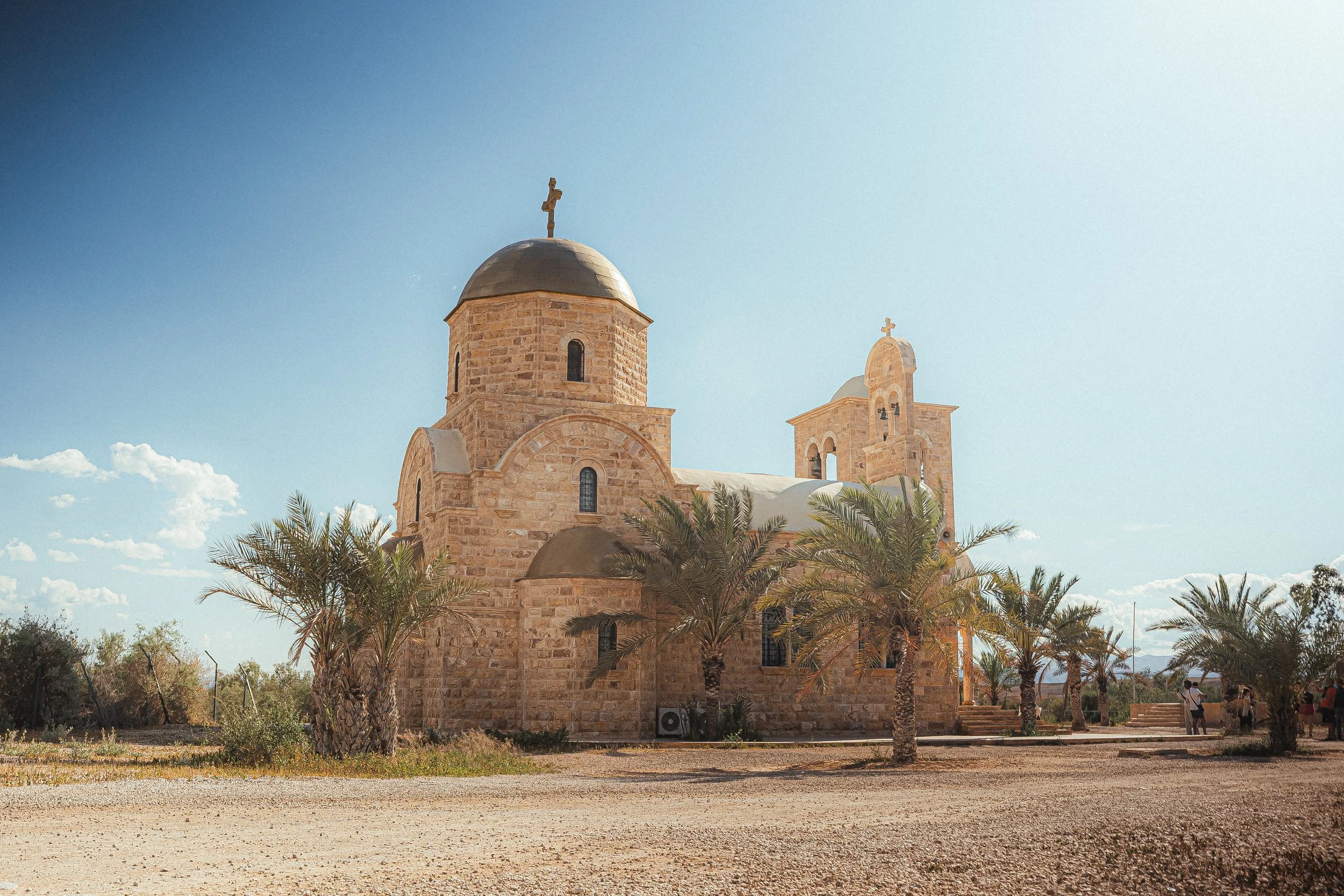 a stone church with domes and a cross on top, surrounded by palm trees and a dirt pathway under a clear blue sky