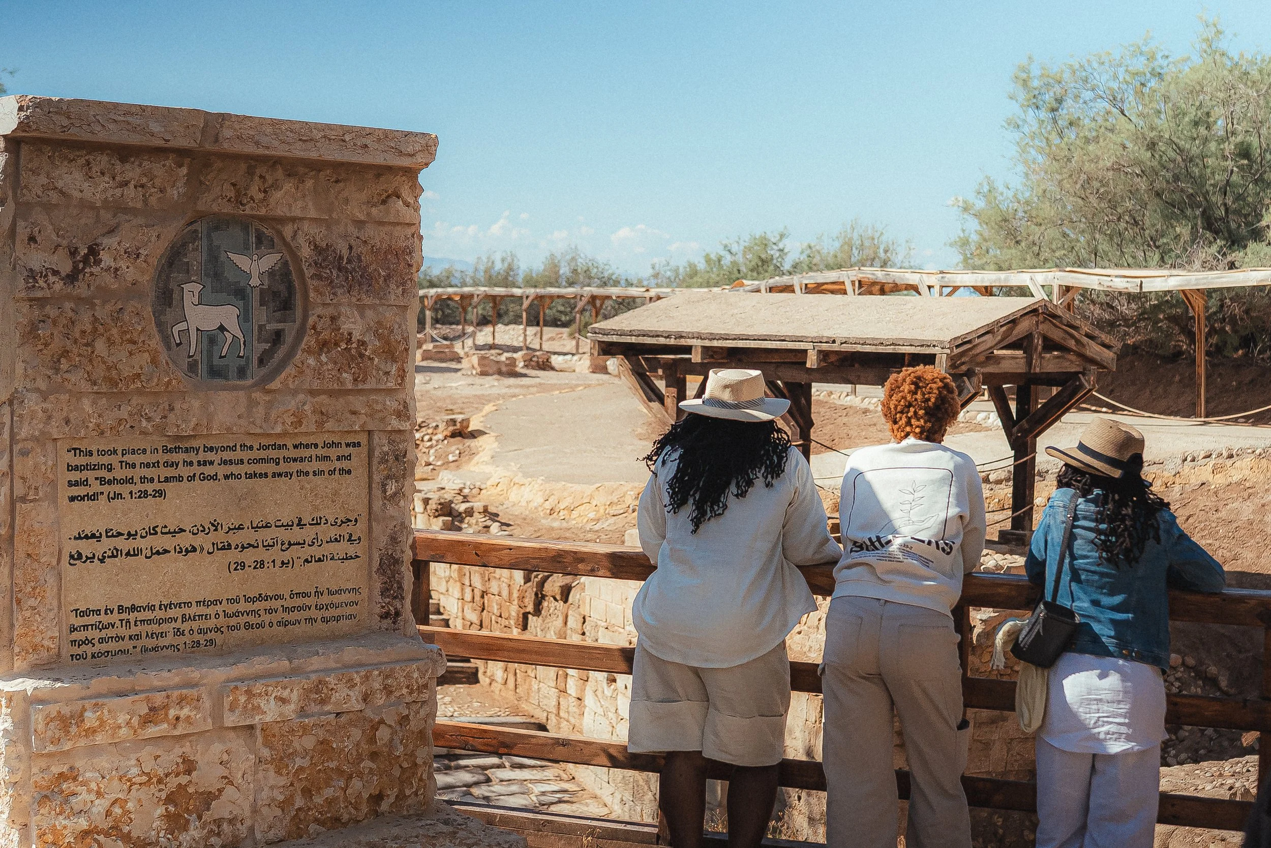 Three people with hats standing by a wooden railing at an archaeological site, looking away from the camera. There is a stone marker with inscriptions and symbols in the foreground on the left side of the image. The background shows ruins, trees, and
