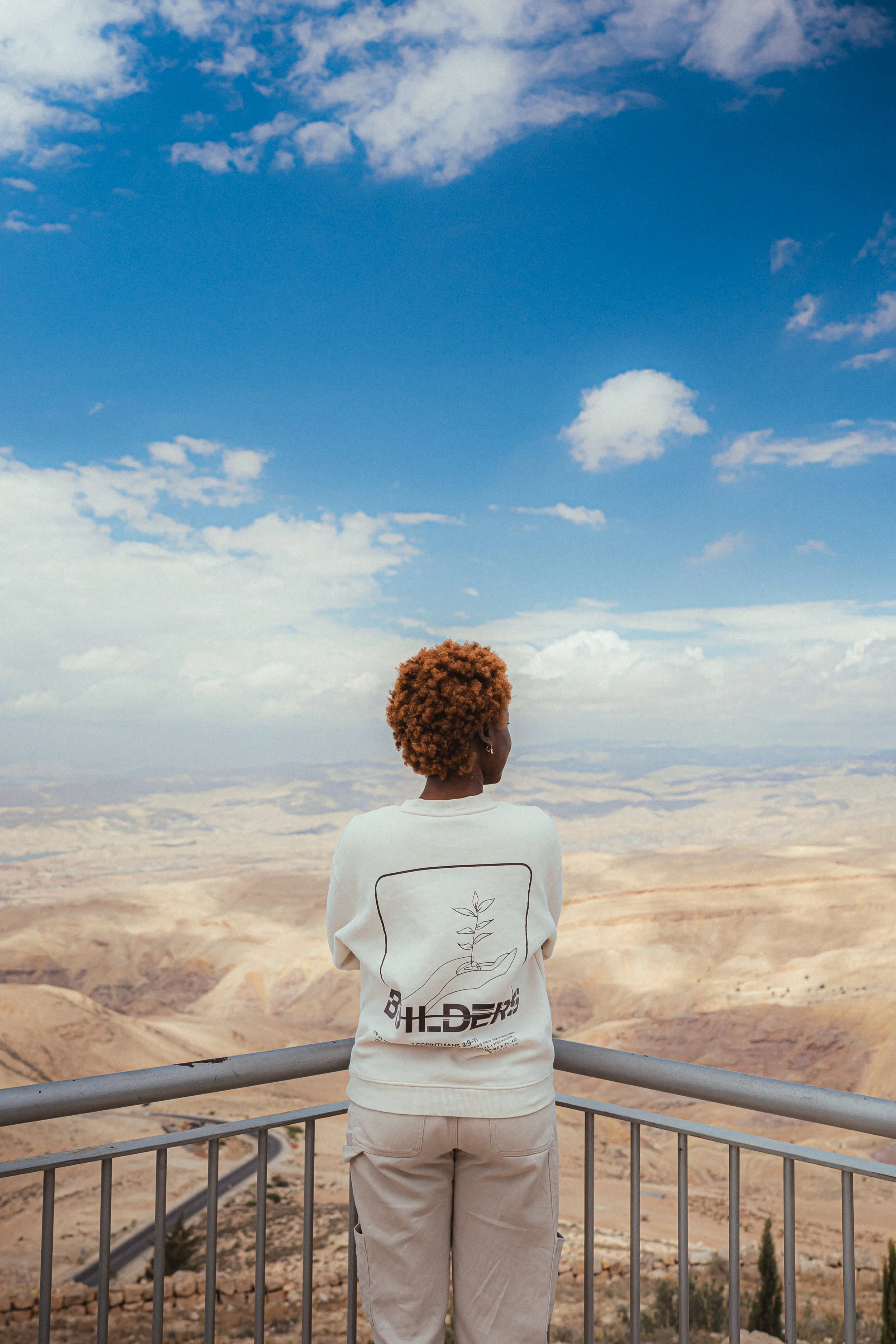Person with short curly hair standing on a viewing platform overlooking a desert landscape, under a partly cloudy blue sky.