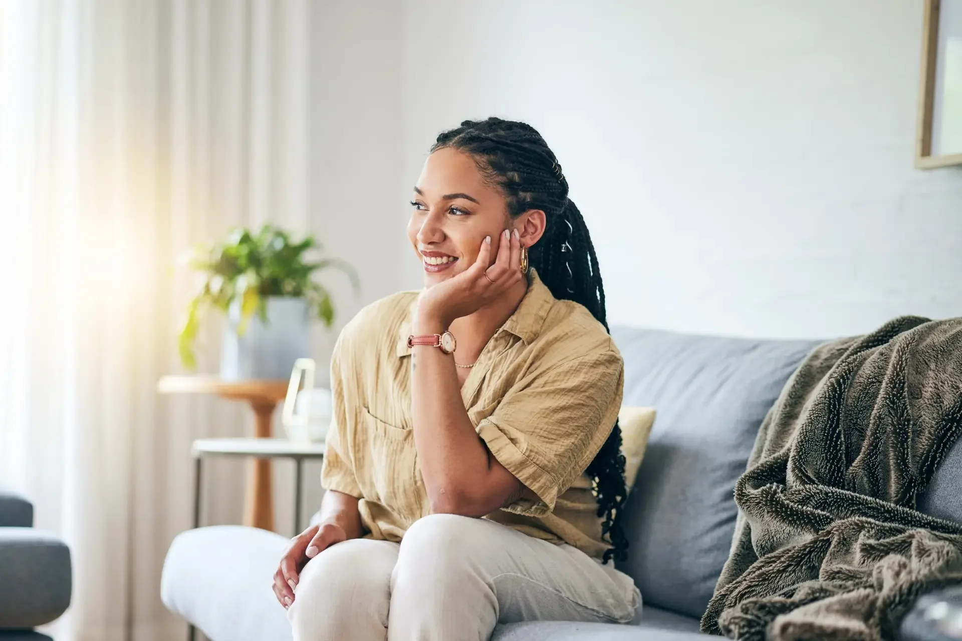 A woman with long braided hair sitting on a gray sofa, smiling, in a well-lit living room with a plant and window in the background.