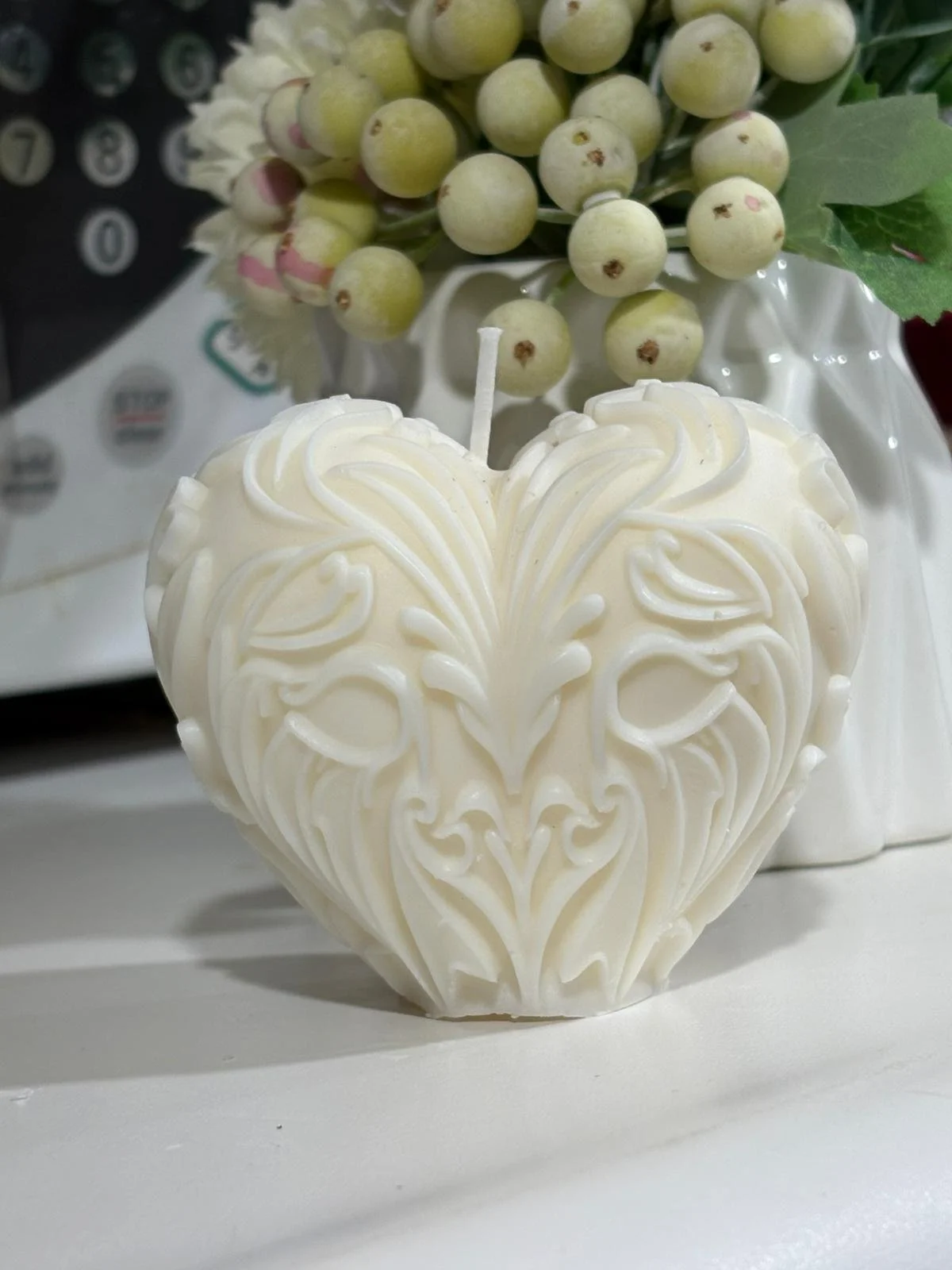 A heart-shaped white candle with ornate patterns, placed on a white surface with a floral arrangement of green berries and small white flowers in the background.