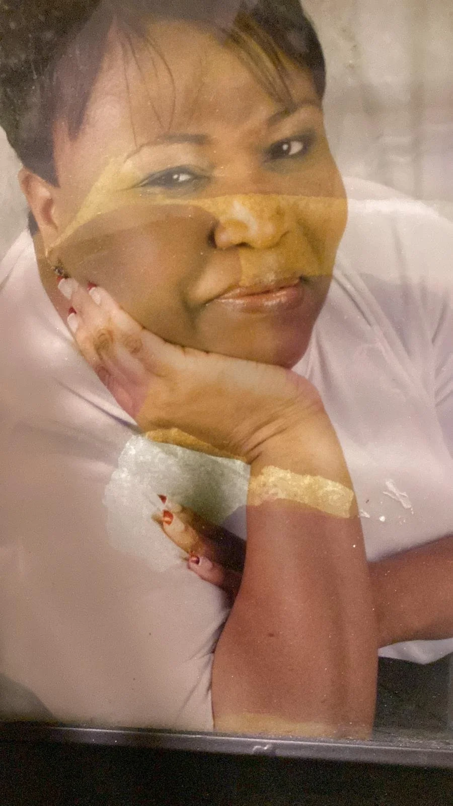 Close-up of a woman resting her chin on her hand, smiling softly, with a somewhat reflective glass or surface overlay creating a layered effect.