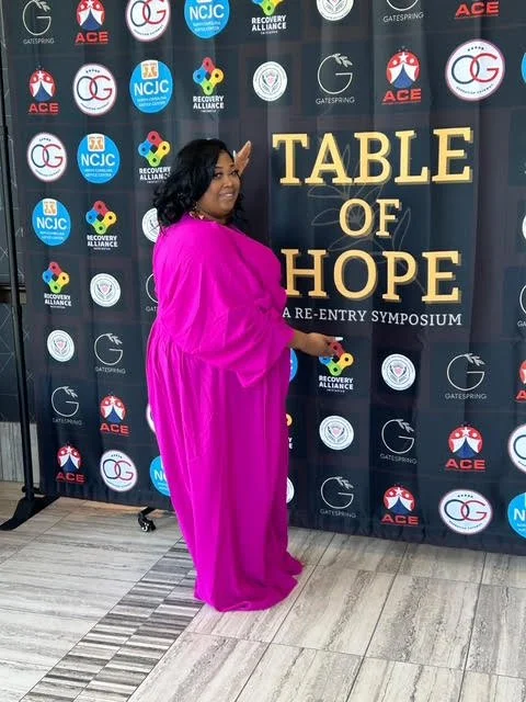 Woman in a bright pink dress standing in front of a black backdrop with various logos and the words "Table of Hope" in gold letters, at a pre-entry symposium.
