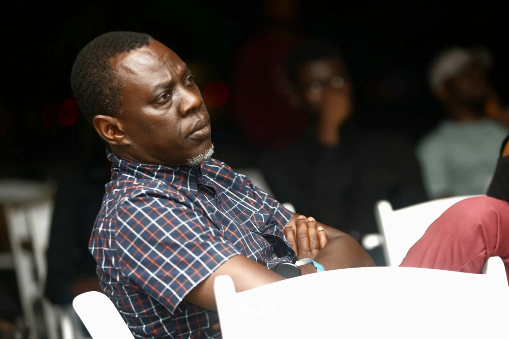 A man with crossed arms sitting in a dark room, wearing a checkered shirt, attentively listening at an event.