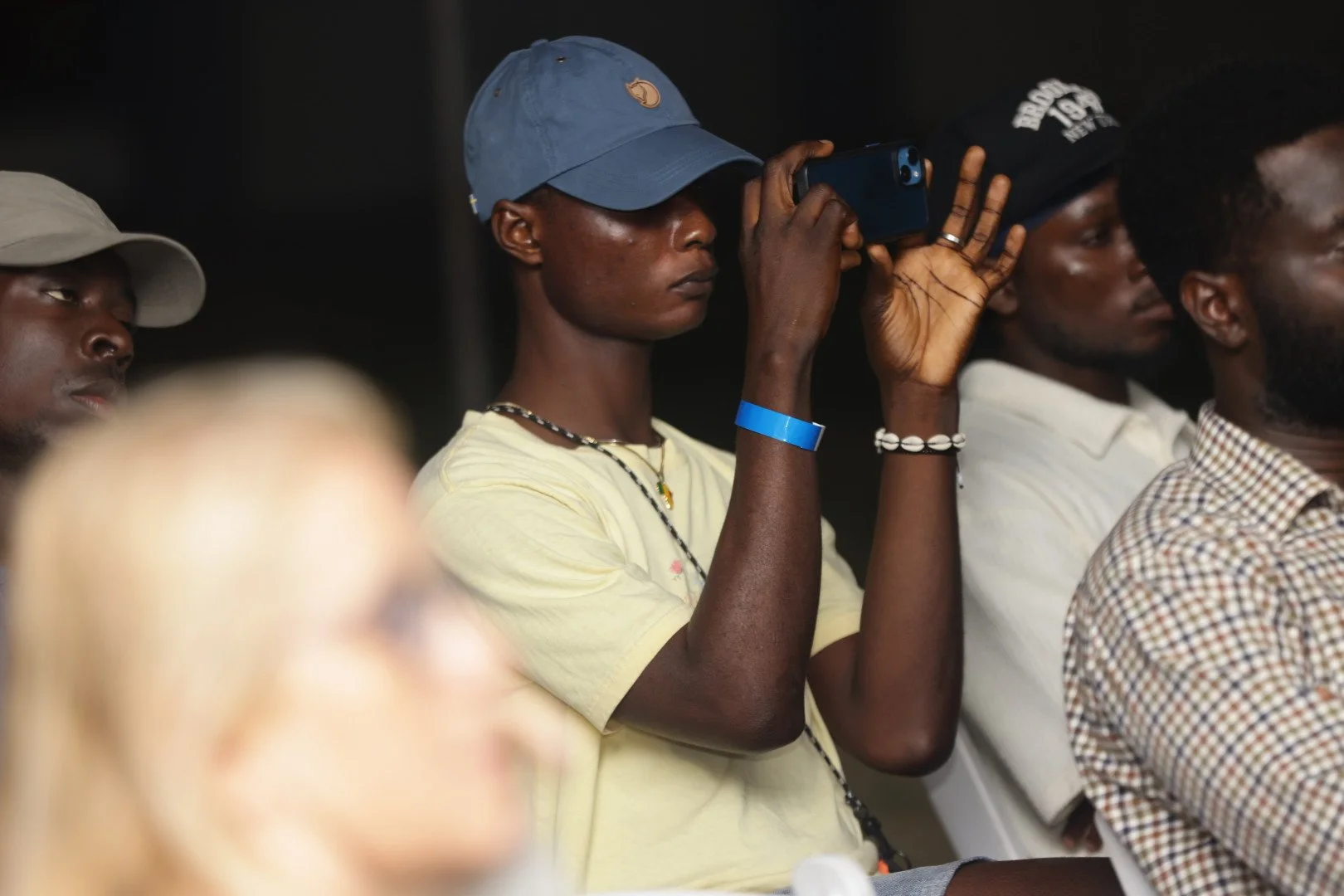 A person wearing a blue cap, yellow shirt, and jewelry is holding a smartphone in a vertical position, taking a photo or video. They are seated among other people attending an event or gathering.
