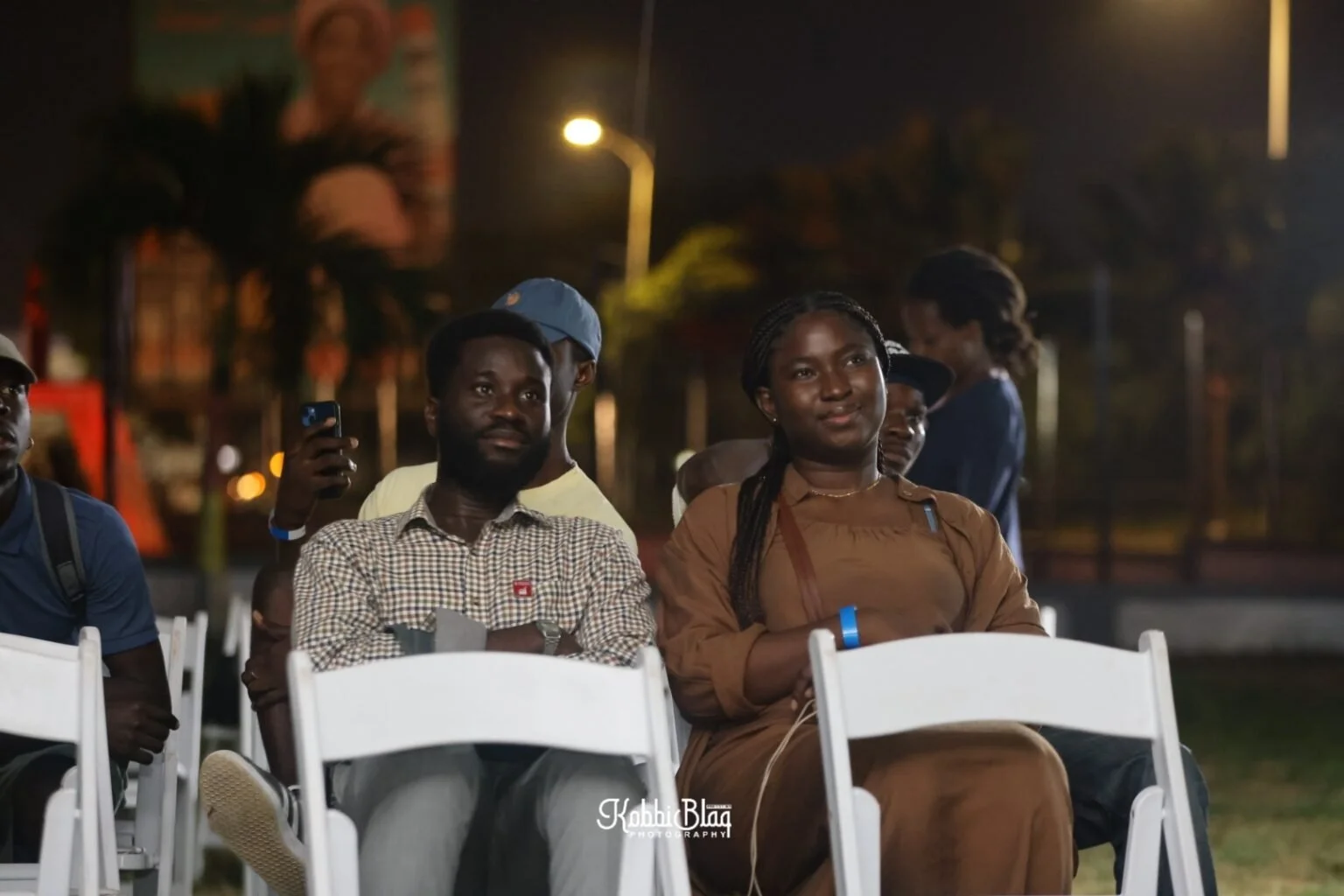 People sitting on white chairs outdoors at night, listening attentively, with some looking at a speaker or presentation, and others taking photos or videos.