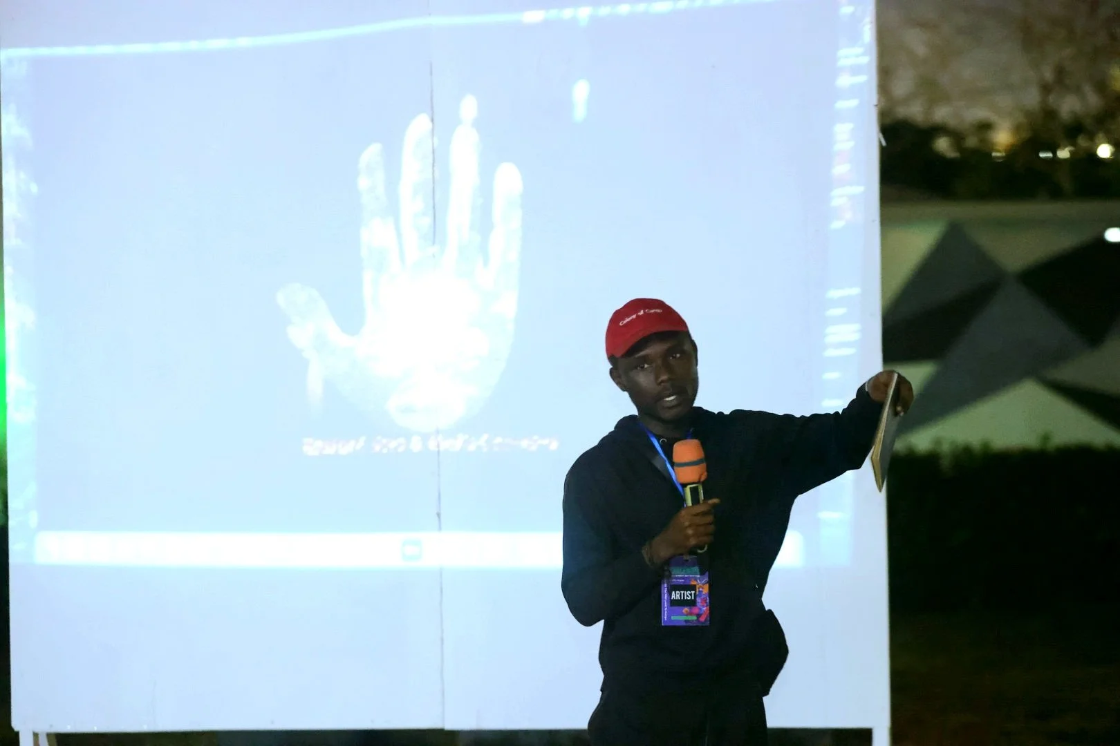 A young man wearing a red cap and black hoodie presenting outdoors at night, holding a microphone in one hand and a paper in the other, standing next to a large screen displaying a digital image of a hand and some text.