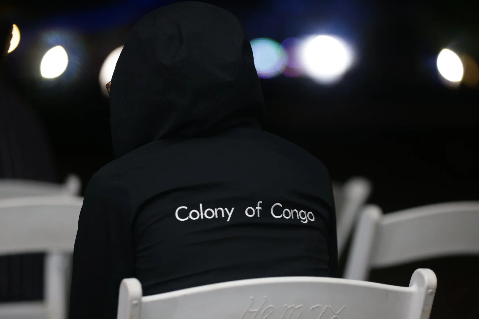 Person wearing a black hoodie with 'Colony of Congo' embroidered on the back, sitting among white chairs at night, with bright lights in the distance.