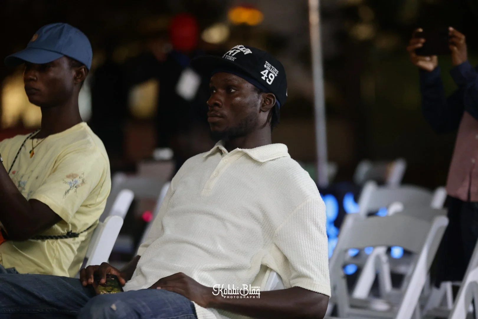 A young man sitting outdoors at night, wearing a white polo shirt and a black cap, looking ahead with a serious expression. There are chairs and other people in the background.