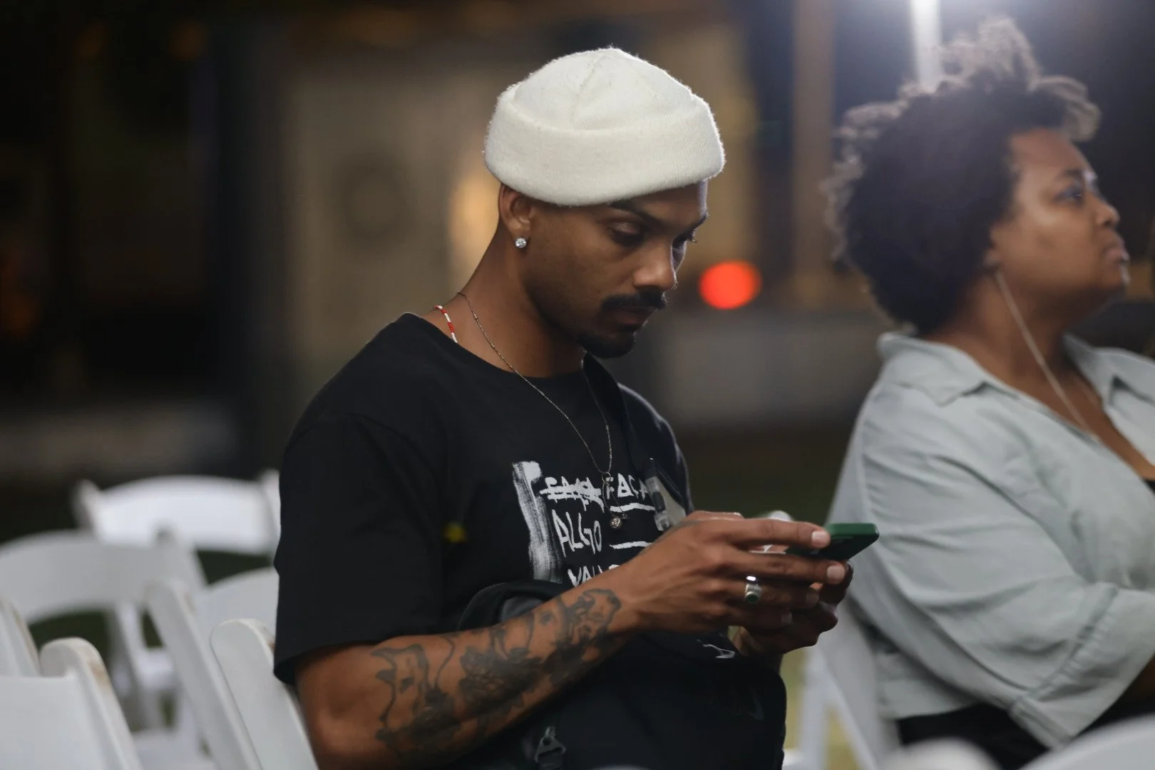 A young man with tattoos on his arms, wearing a white beanie hat, earrings, and a black T-shirt, is sitting on white chairs outdoors at night, looking at his phone. Next to him, an older woman with curly hair and a light-colored shirt is sitting with